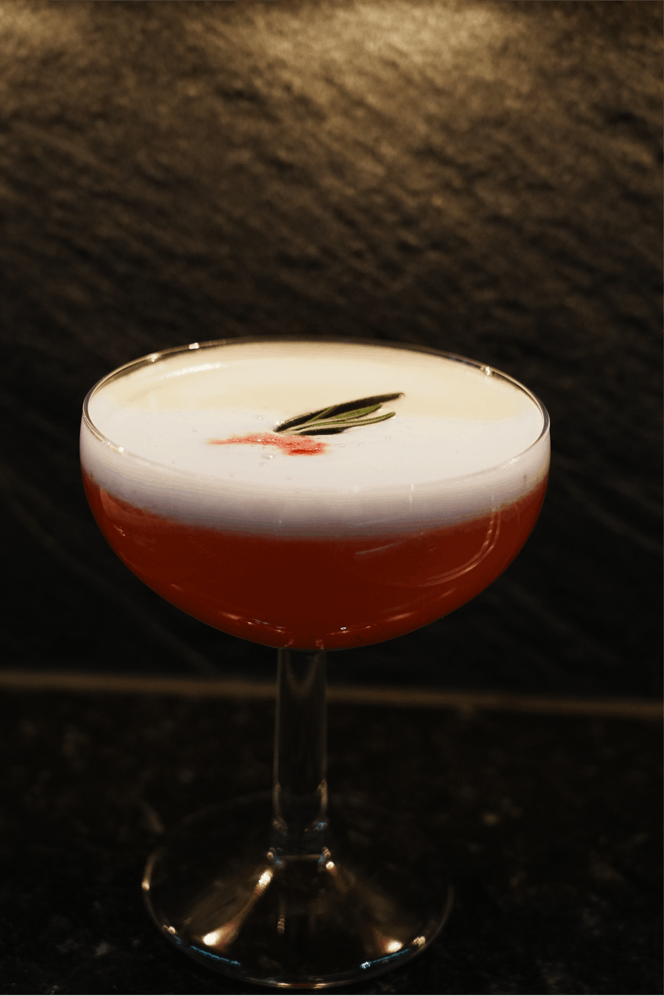 En cocktail i ett cocktailglas med ett lager av skum, dekorerad med en rosmarastjälk och en liten dropp av röd sirap, placerad på en mörk yta mot en stenvägg.