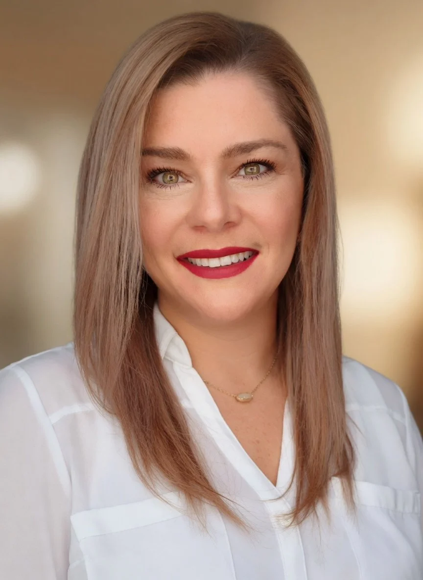 A woman with shoulder-length light brown hair and light skin, wearing a white blouse, red lipstick, and a small necklace, smiling at the camera.