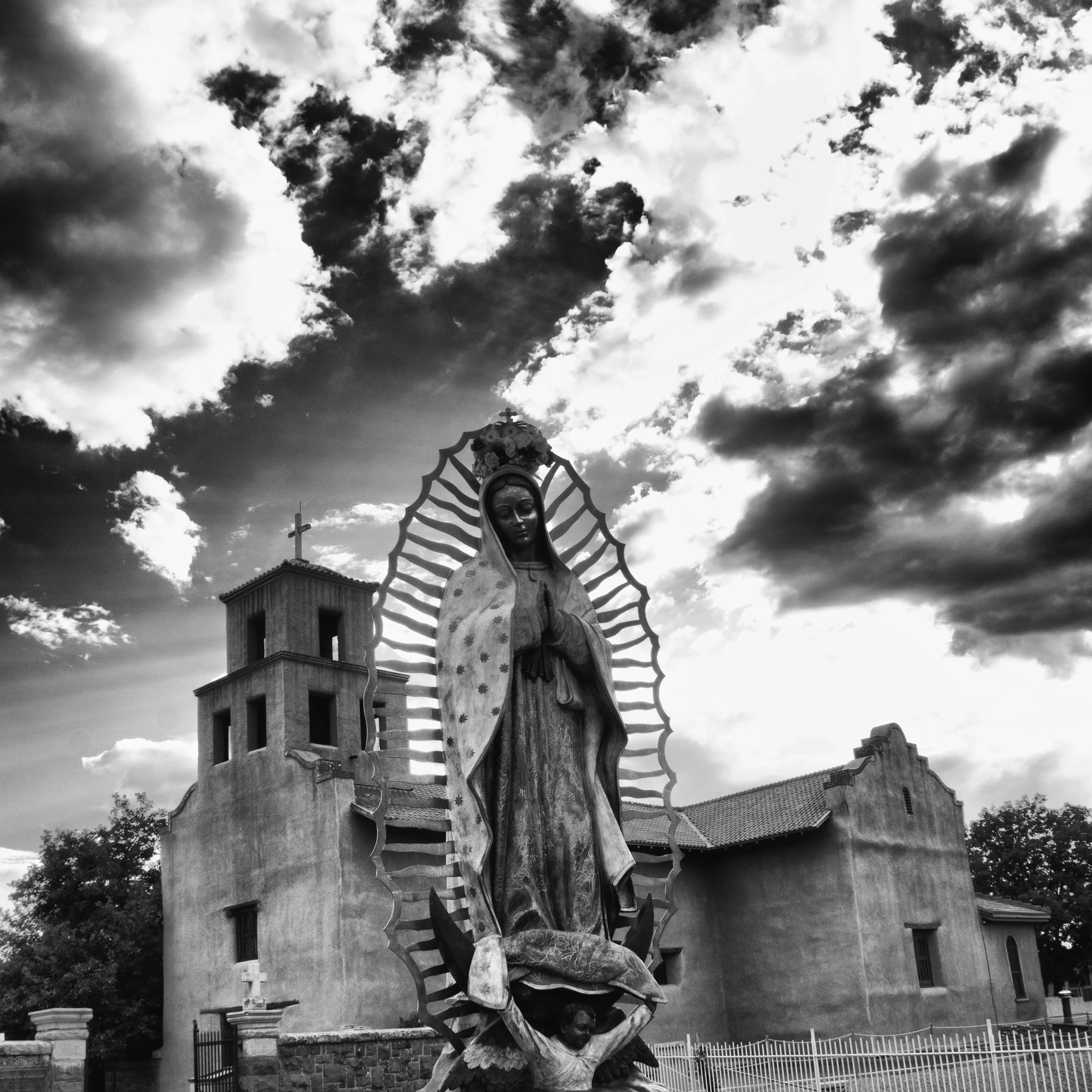 Black and white photo of a religious statue of the Virgin Mary with a floral crown, hands clasped in prayer, standing on a turtle held by a person. A church building with a bell tower and a cross is visible in the background, with cloudy sky overhead.