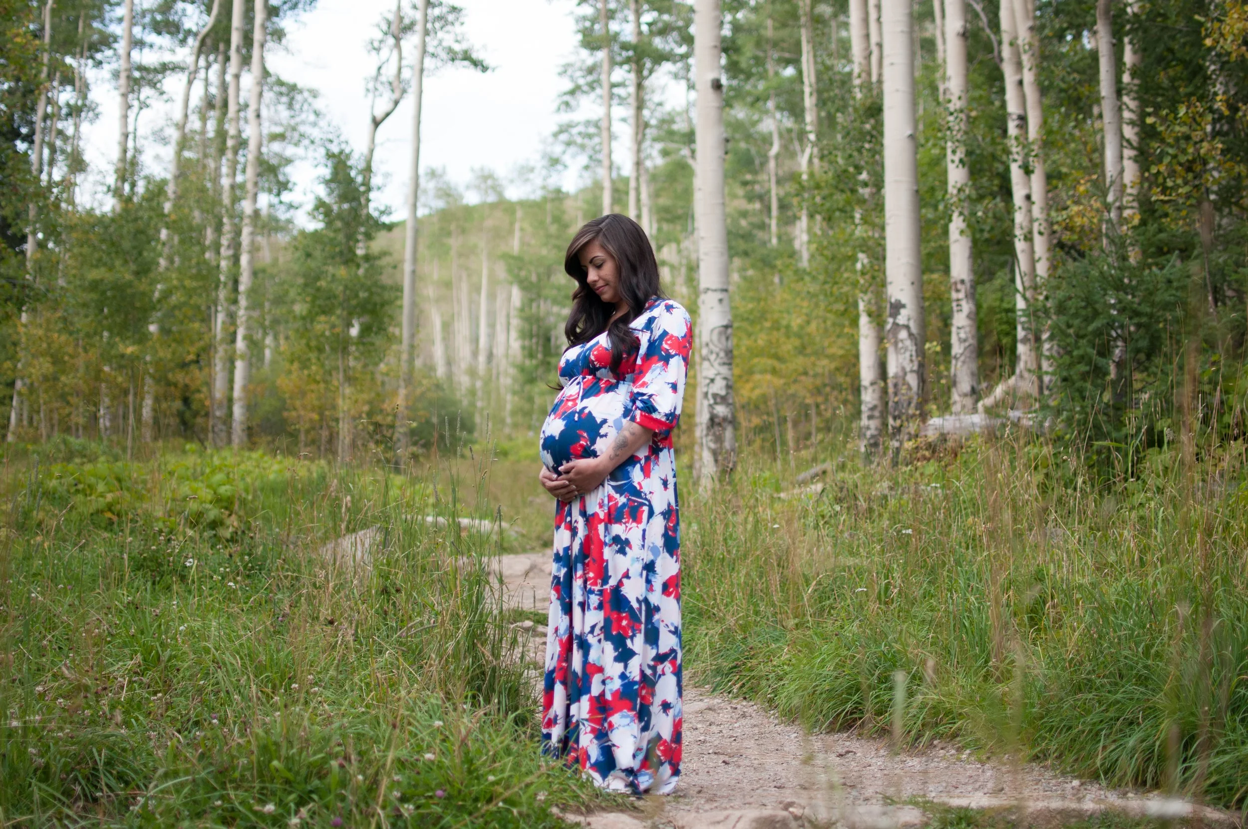 Pregnant woman in a floral dress standing on a forest trail, gently holding her belly and looking down, surrounded by trees and green foliage.