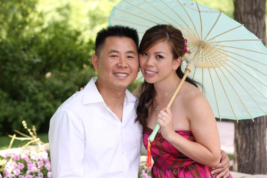A smiling man and woman pose outdoors with a light green parasol, surrounded by trees and pink flowers.