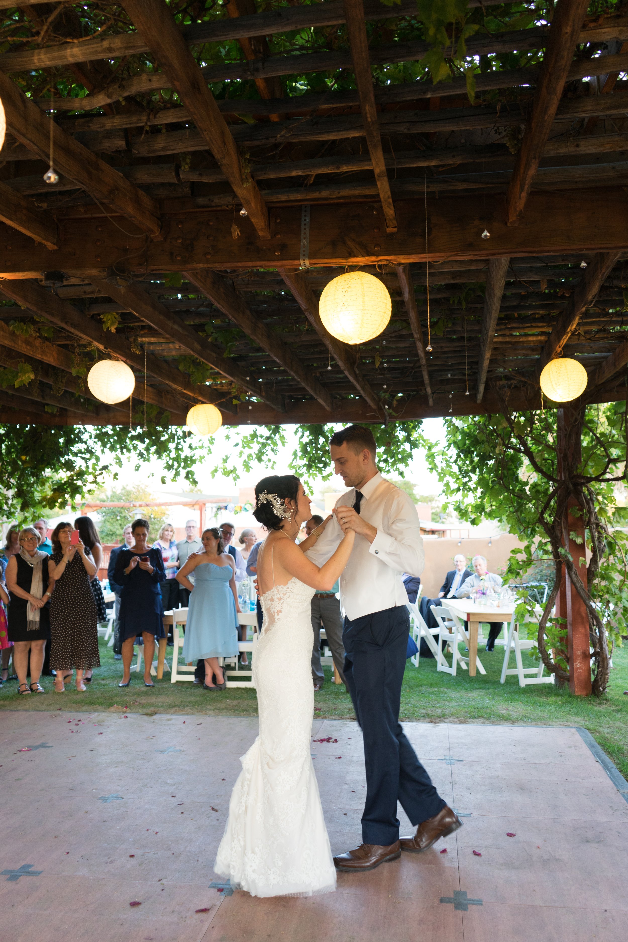 A bride and groom are dancing at their wedding reception under a wooden pergola decorated with hanging paper lanterns, with guests watching in the background.