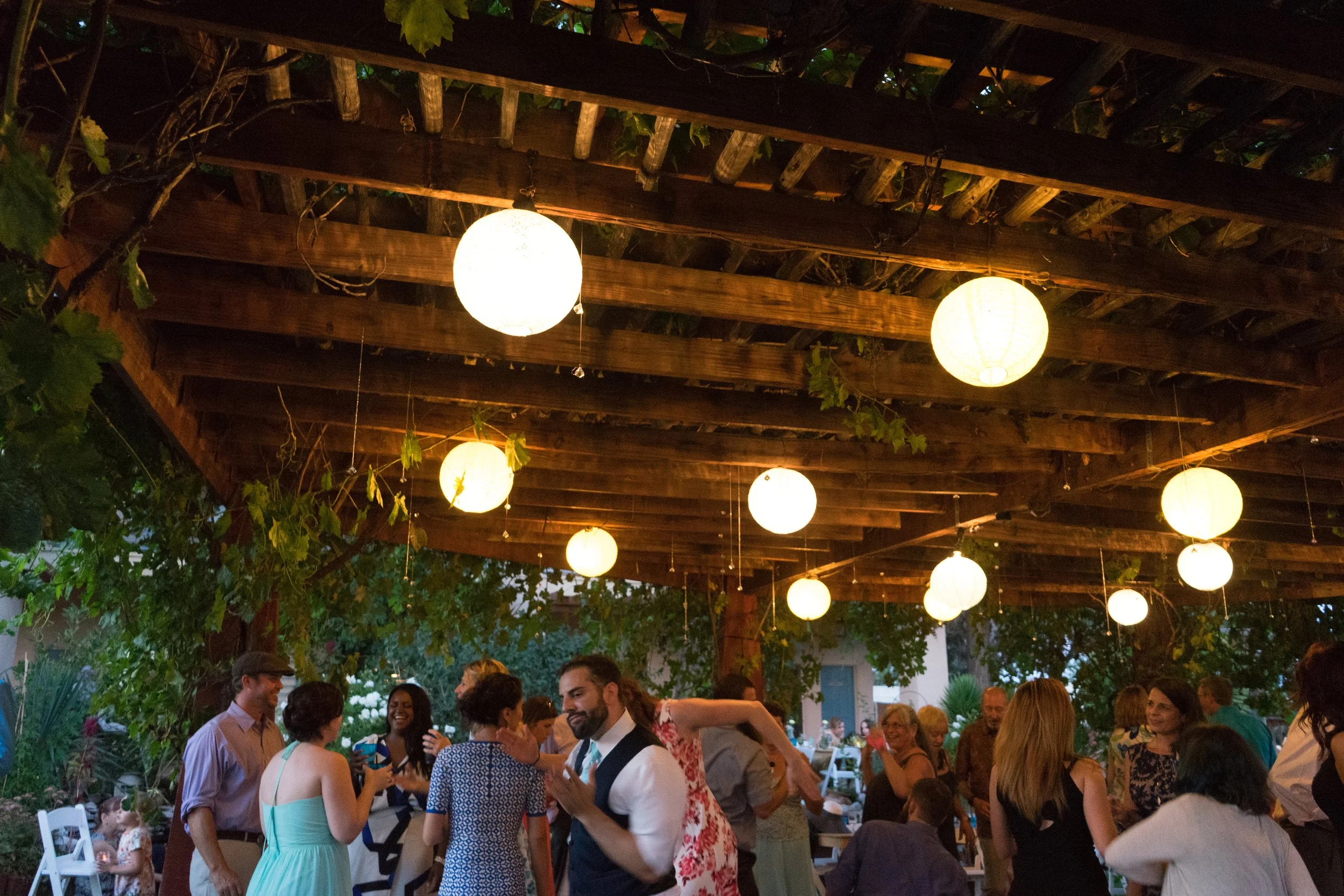 People socializing at an outdoor party under a wooden pergola decorated with hanging paper lanterns at dusk
