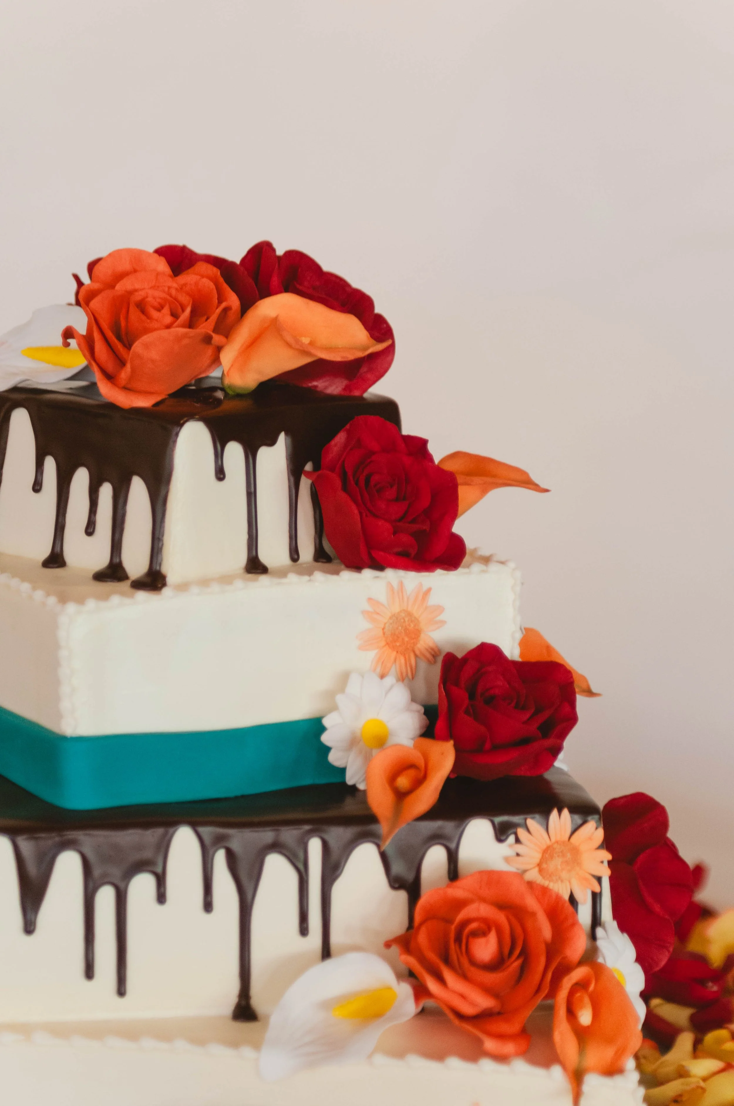 Two-tiered wedding cake decorated with red, orange, and white flowers, topped with chocolate drizzle.