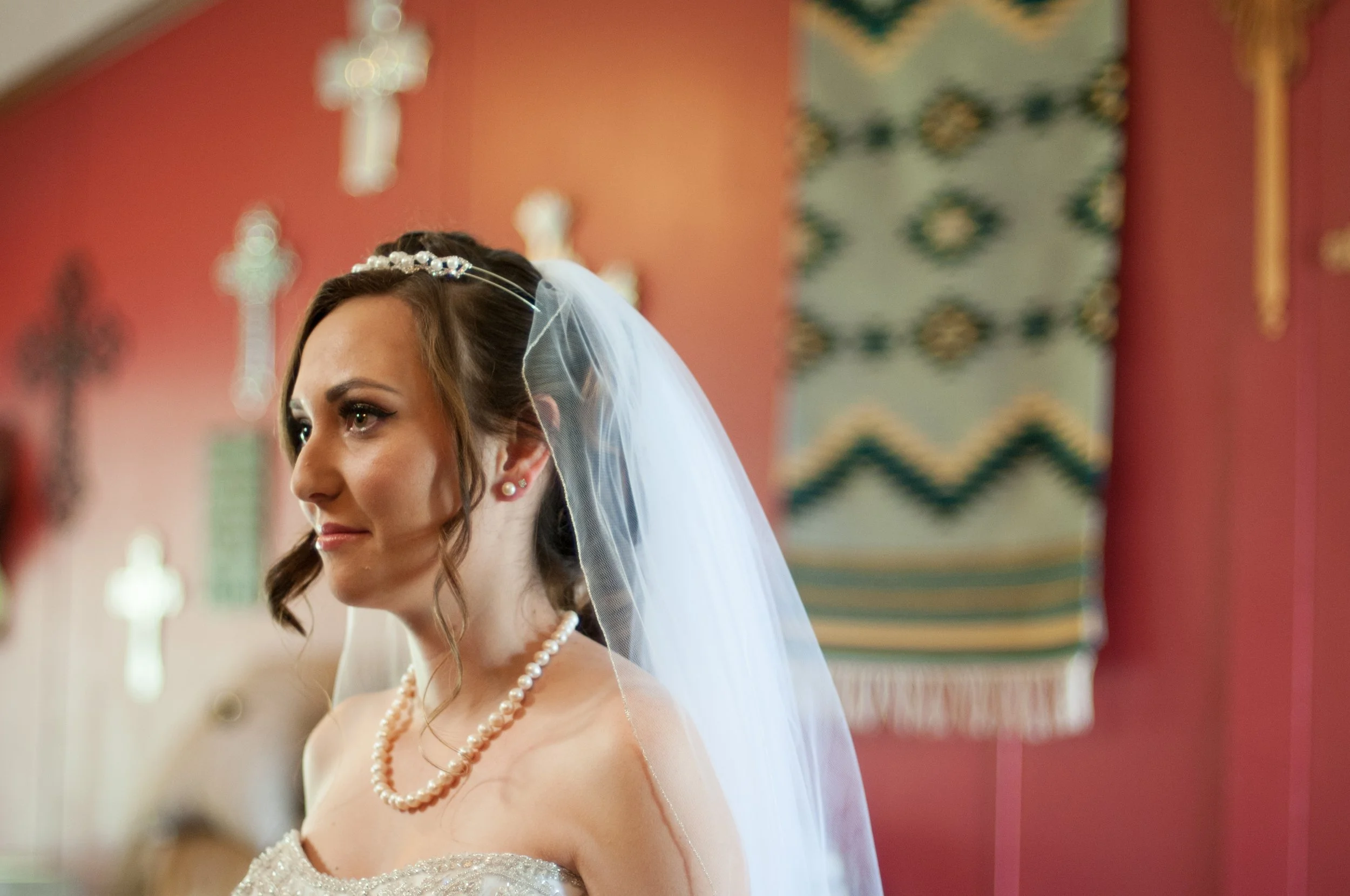 A bride with a veil, pearl necklace, and earrings looking to her left in a church with biblical crosses and a colorful tapestry in the background.