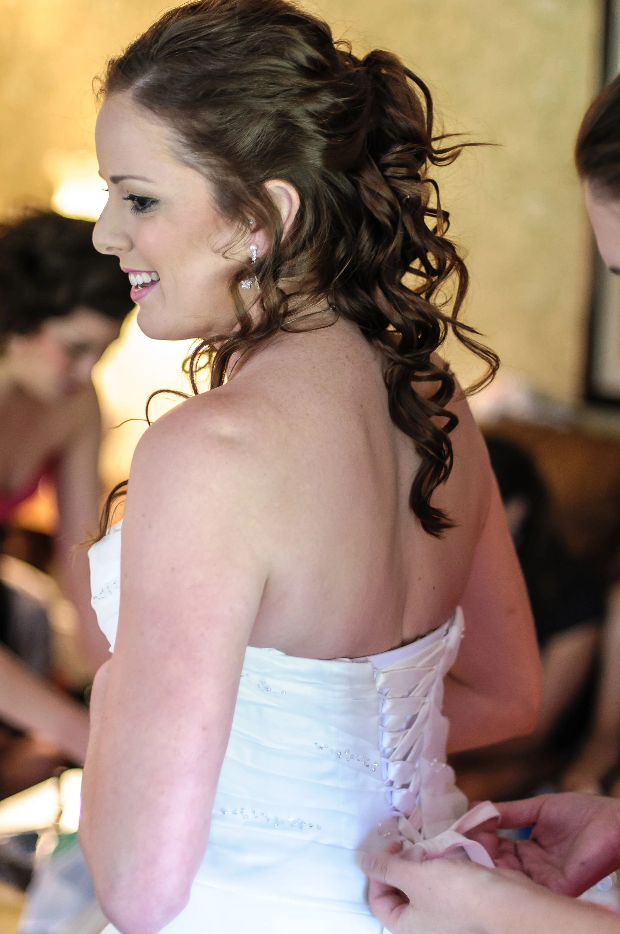 A woman in a white wedding dress being helped into her wedding gown, with dark, curly hair styled elegantly, in a softly lit room.