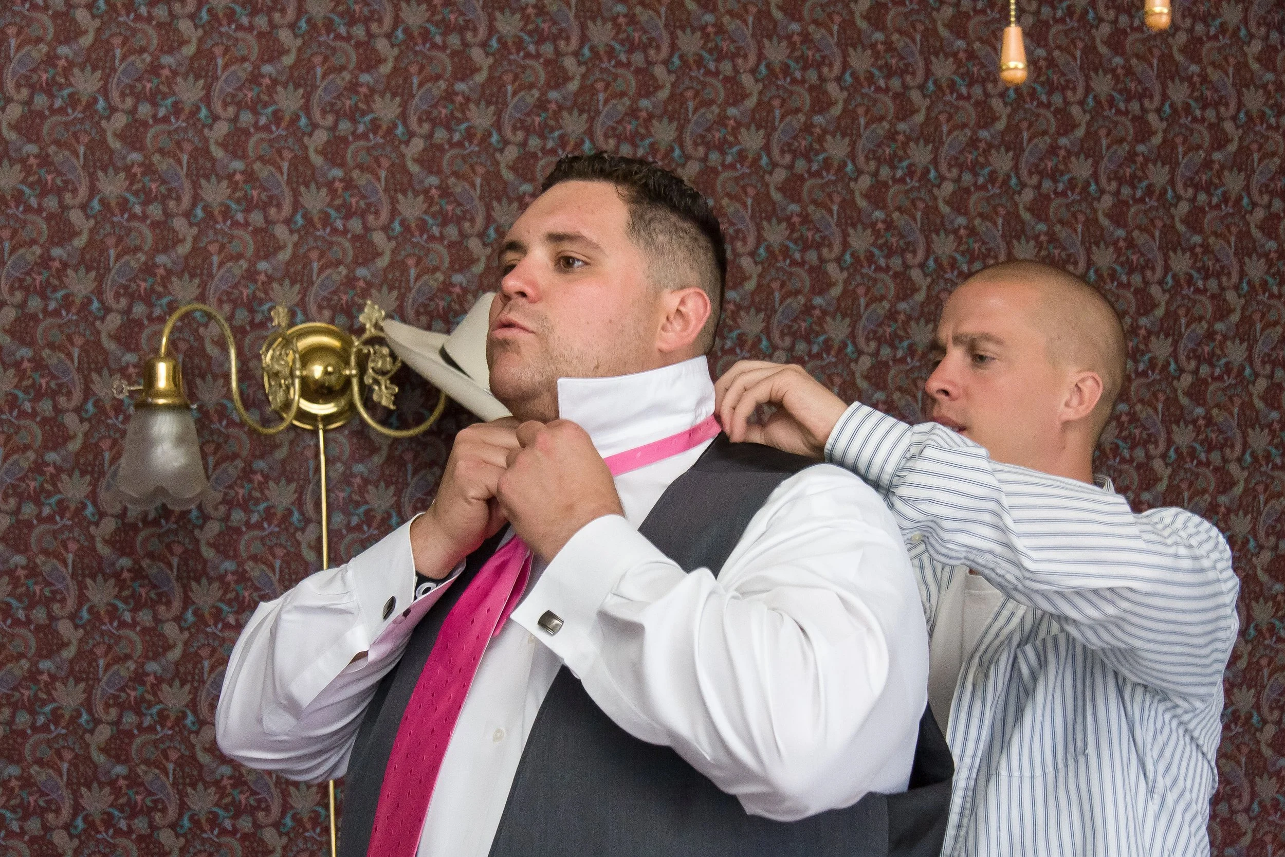 Two men in formal attire, one adjusting the other's collar in a room with ornate wallpaper and a wall-mounted lamp.