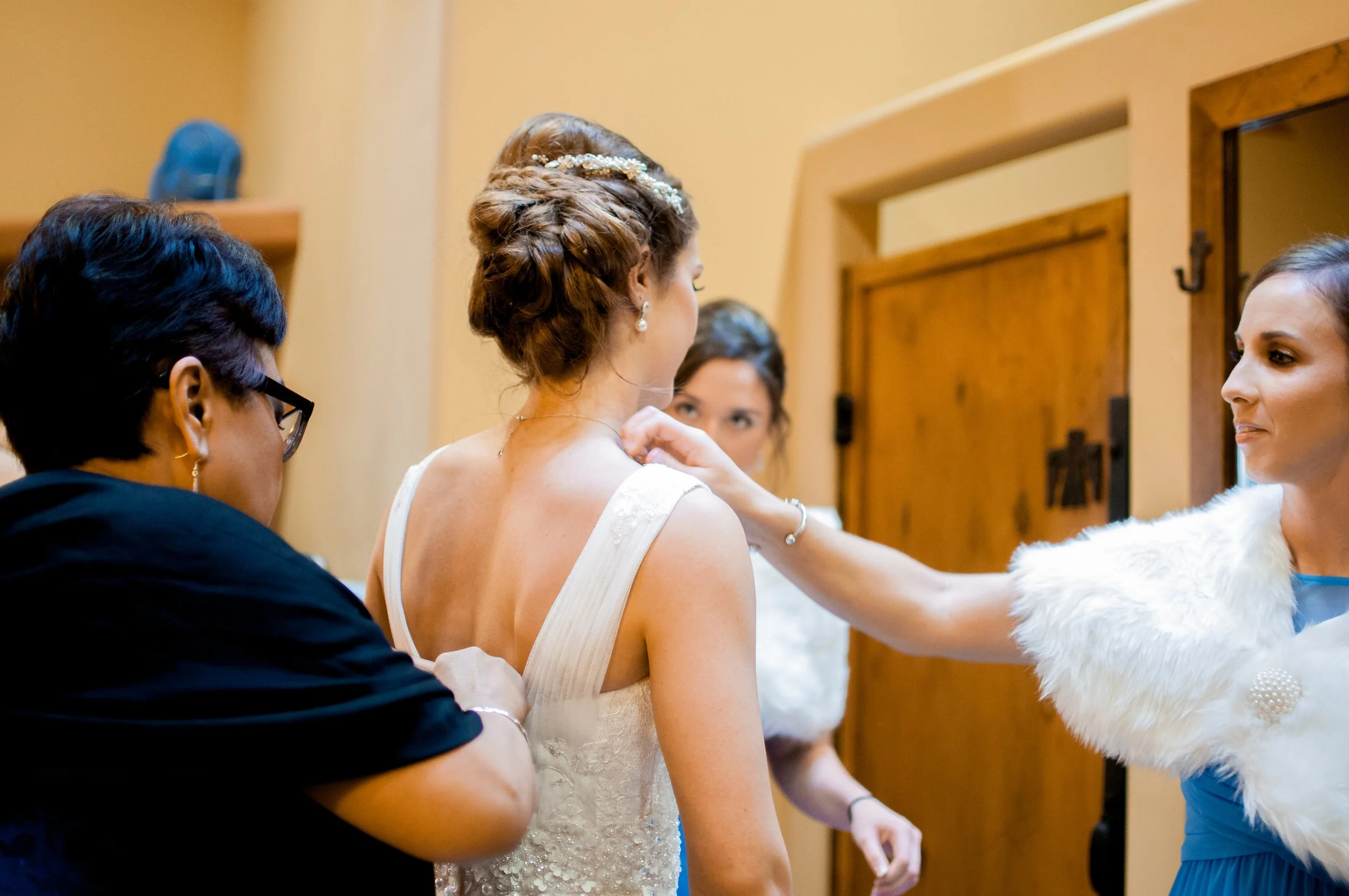 Four women preparing a bride for her wedding, with one woman adjusting the bride's dress and others observing.