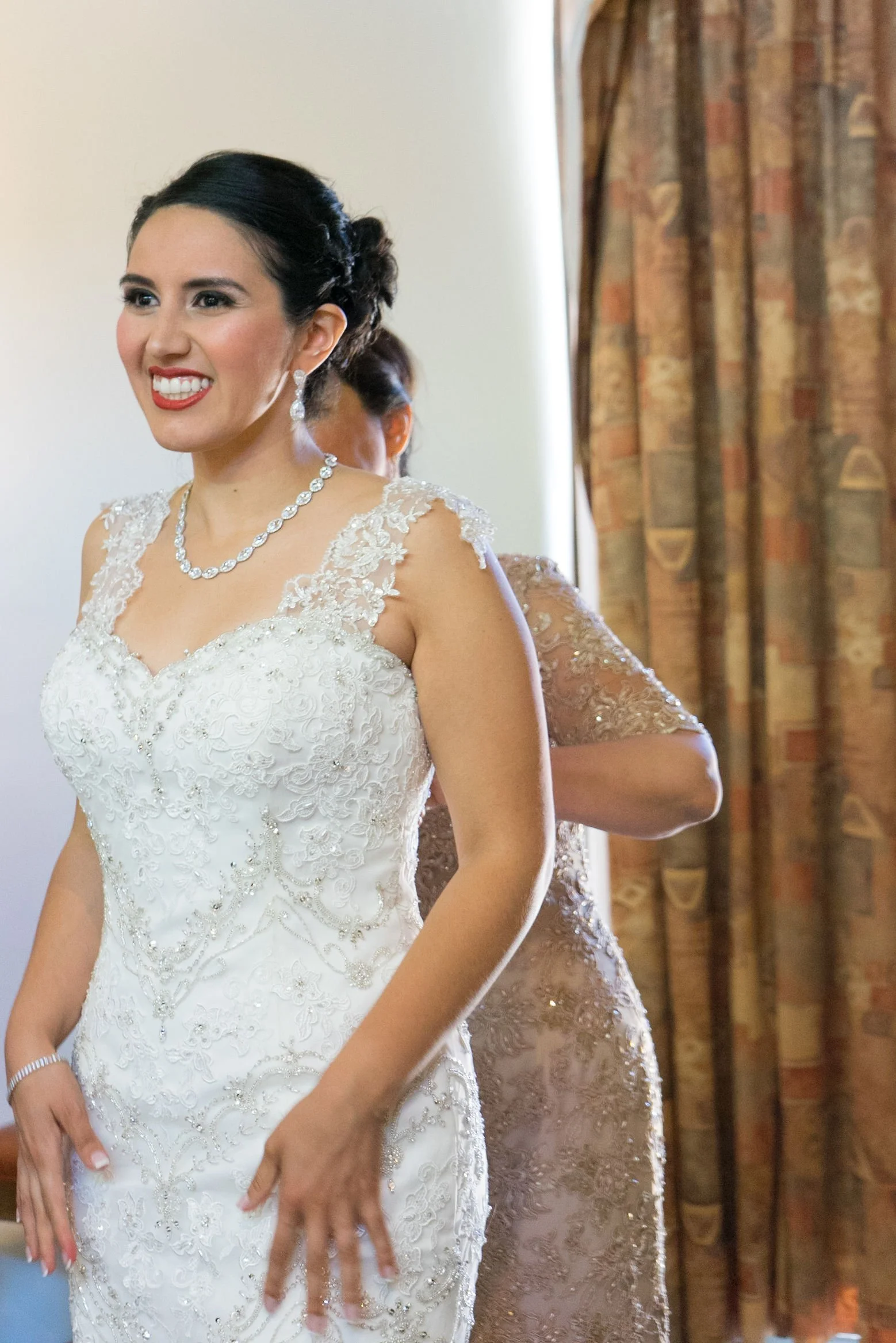A bride wearing a white wedding dress decorated with lace and beading, with earrings, necklace, and bracelet, smiling as someone helps her with her dress in a room with patterned curtains.
