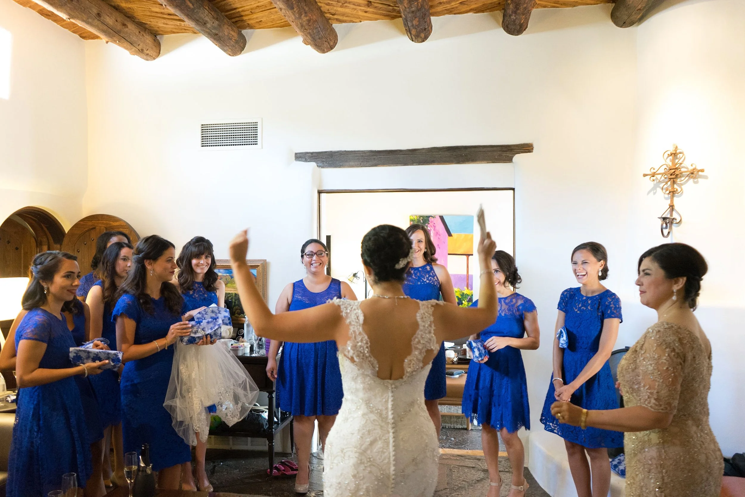 Bride in a white wedding dress with lace details surrounded by seven bridesmaids in matching blue dresses, in a room with white walls and wooden beams, as part of a wedding celebration.