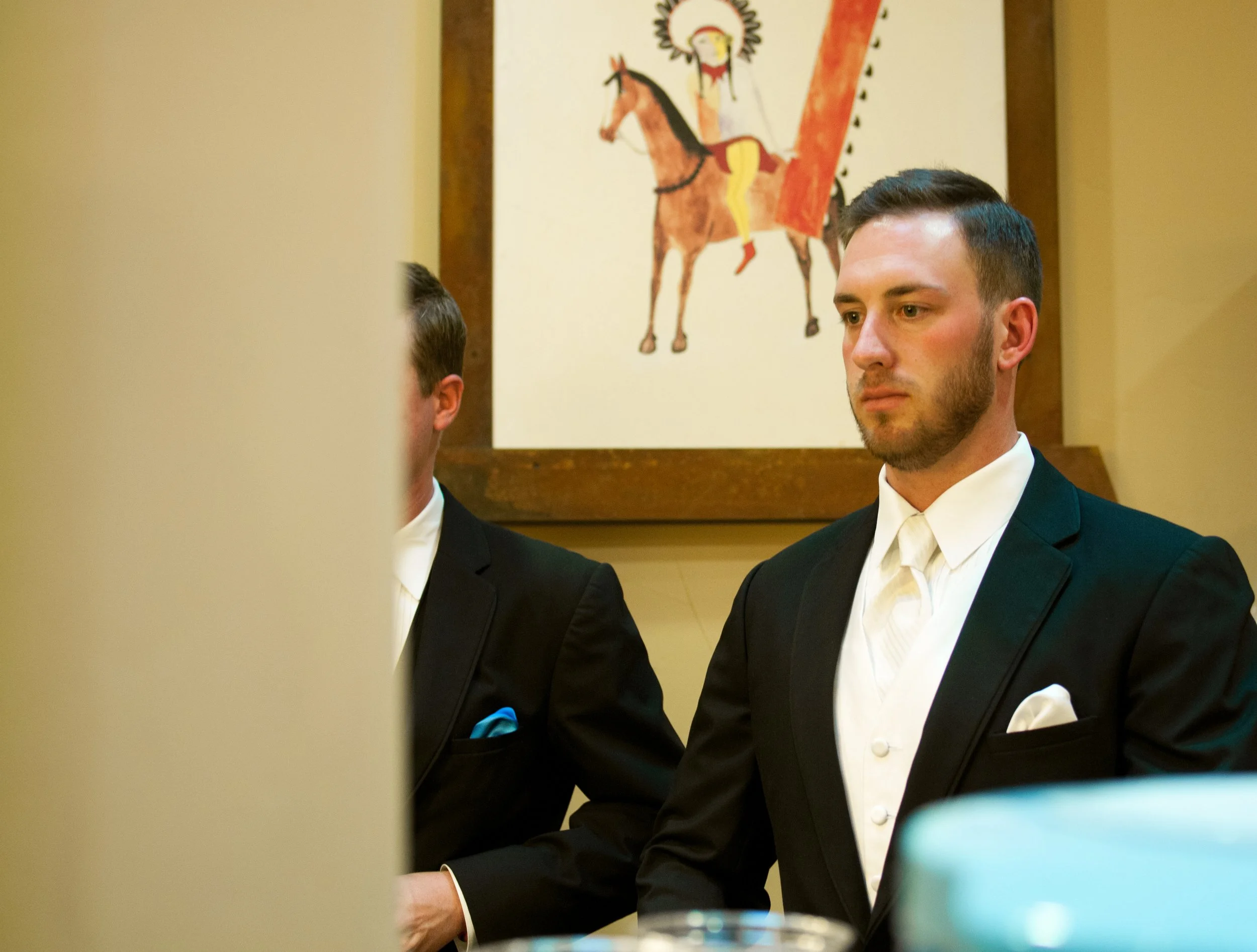 A man in a black tuxedo with a white vest and bow tie is sitting at a formal event. There is a partially visible person to his left and a painting of a person riding a horse in the background.