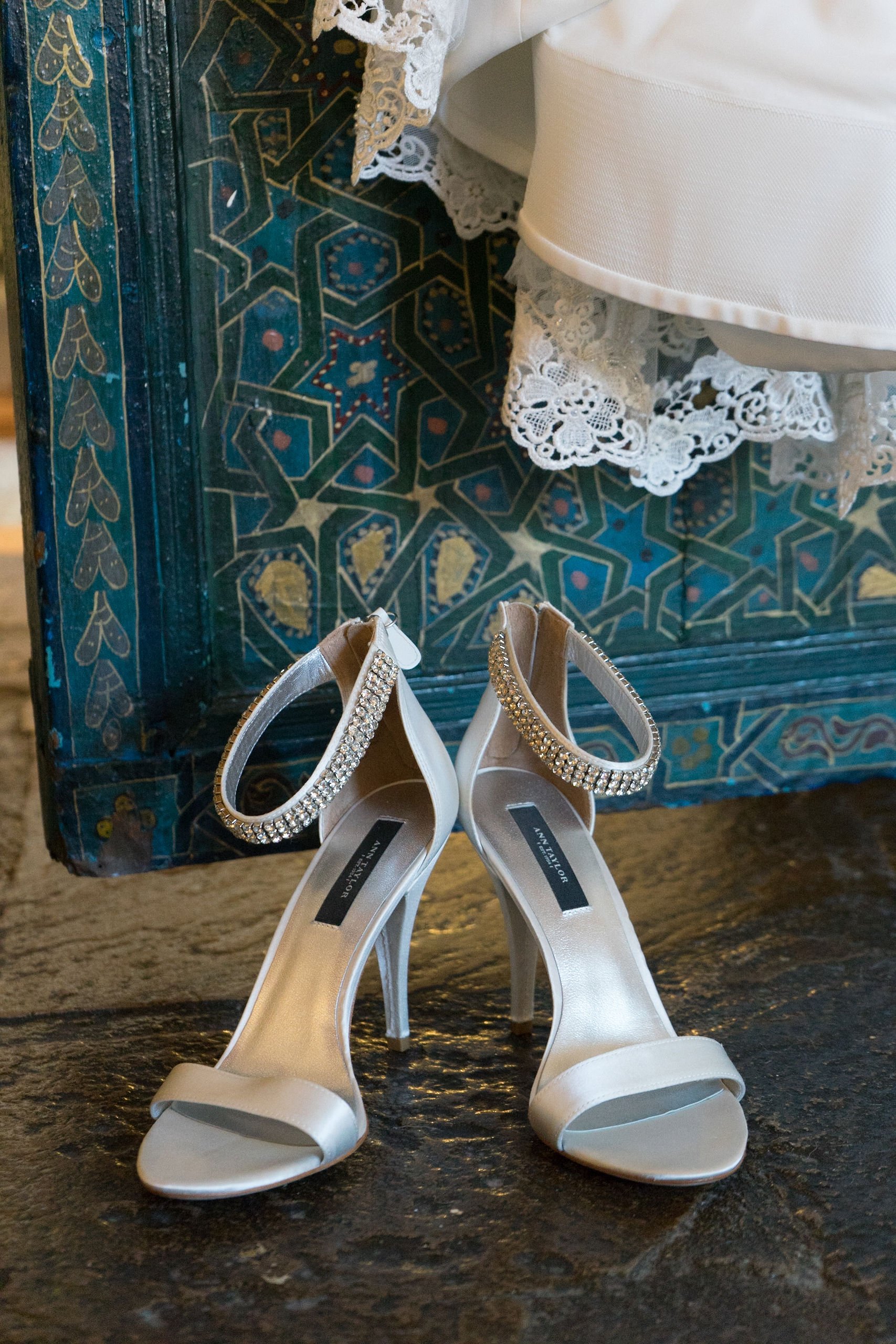 A pair of white high-heeled shoes with rhinestone ankle straps, placed on a dark textured floor. A decorative, colorful, and intricately patterned blue and gold furniture or floor is in the background, alongside a white dress with lace details.
