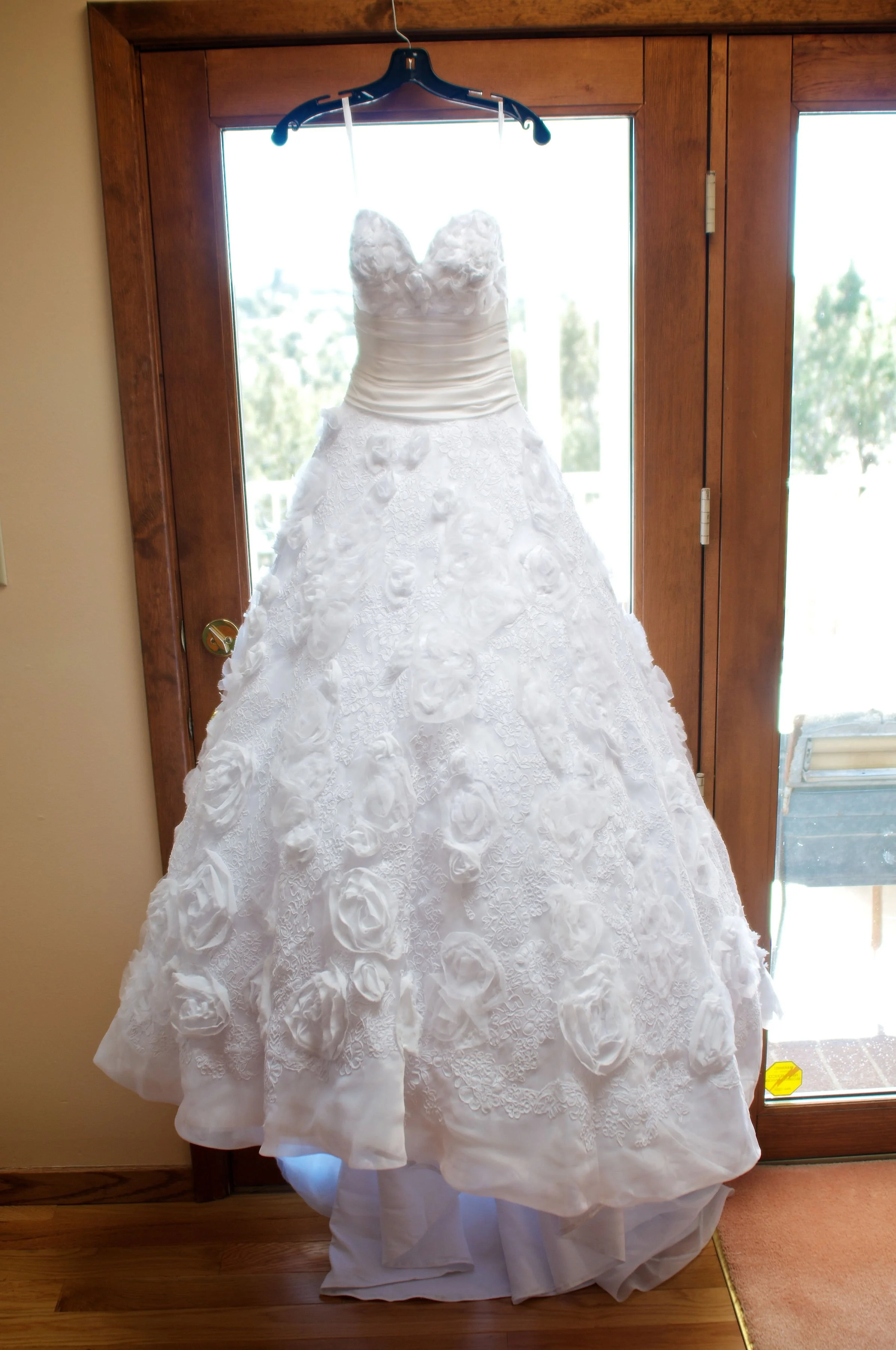 White wedding dress with floral lace and rosette embroidery hanging on a black hanger in front of a glass door with wooden frame.