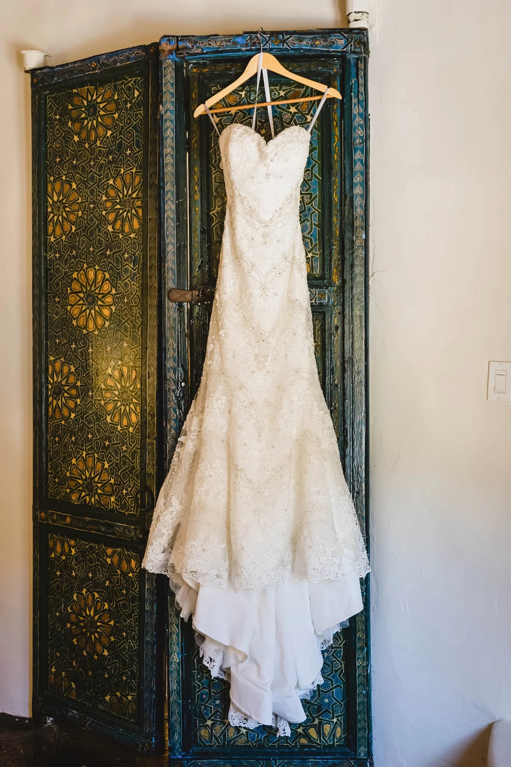 White wedding dress hanging on a wooden hanger in front of a decorative, colorful folding screen.