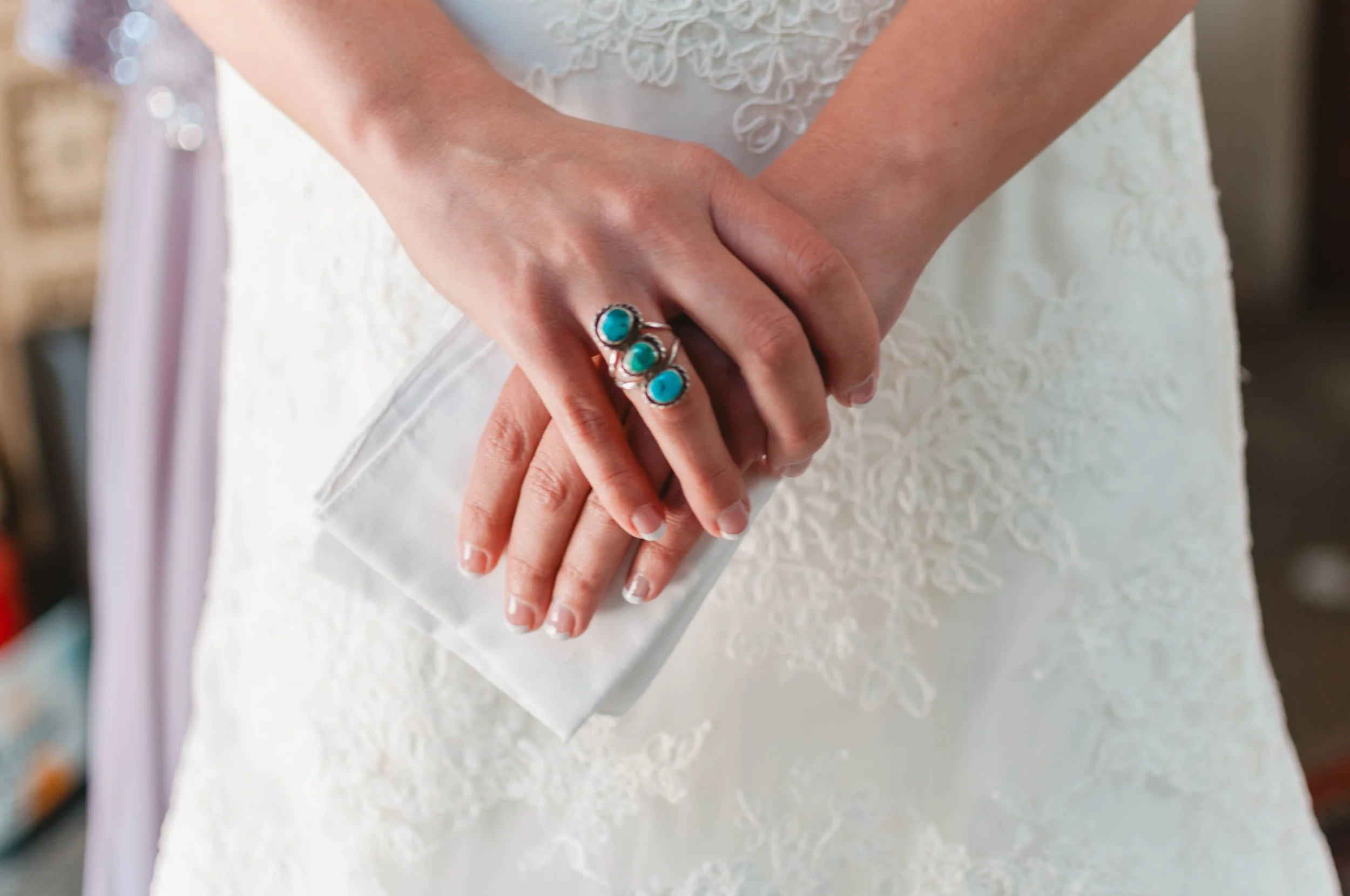 A person in a white lace dress is holding a white clutch purse with hands adorned with a silver ring featuring three turquoise stones.