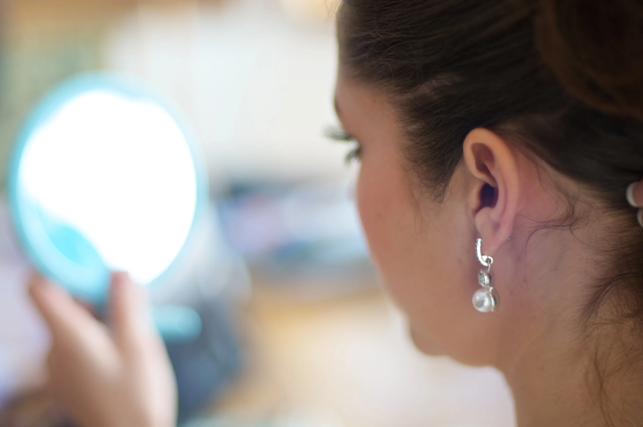 A woman with brown hair and earrings looking into a bright, round mirror.