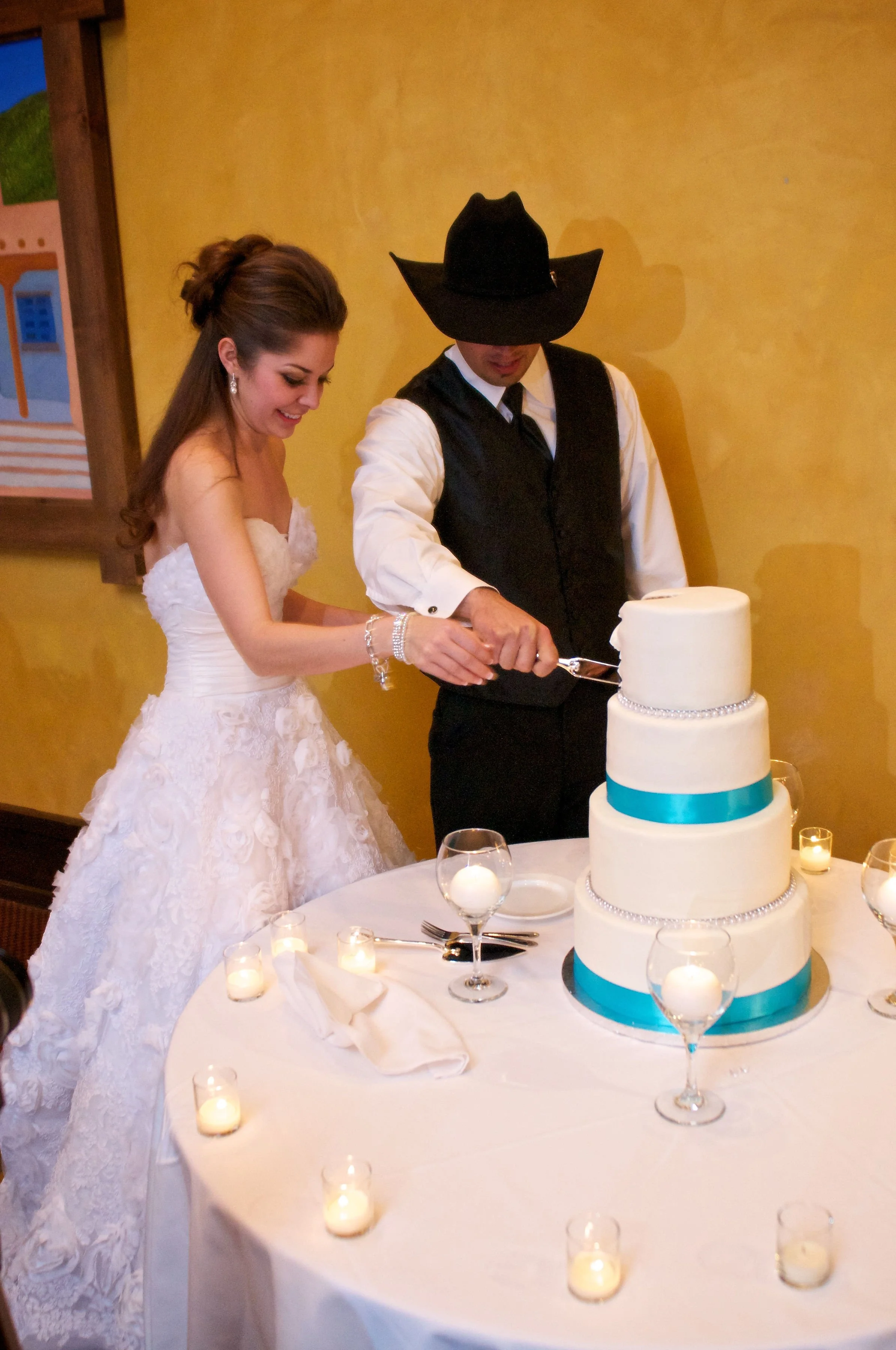 Bride and groom cutting wedding cake at reception, surrounded by lit candles on a round table.