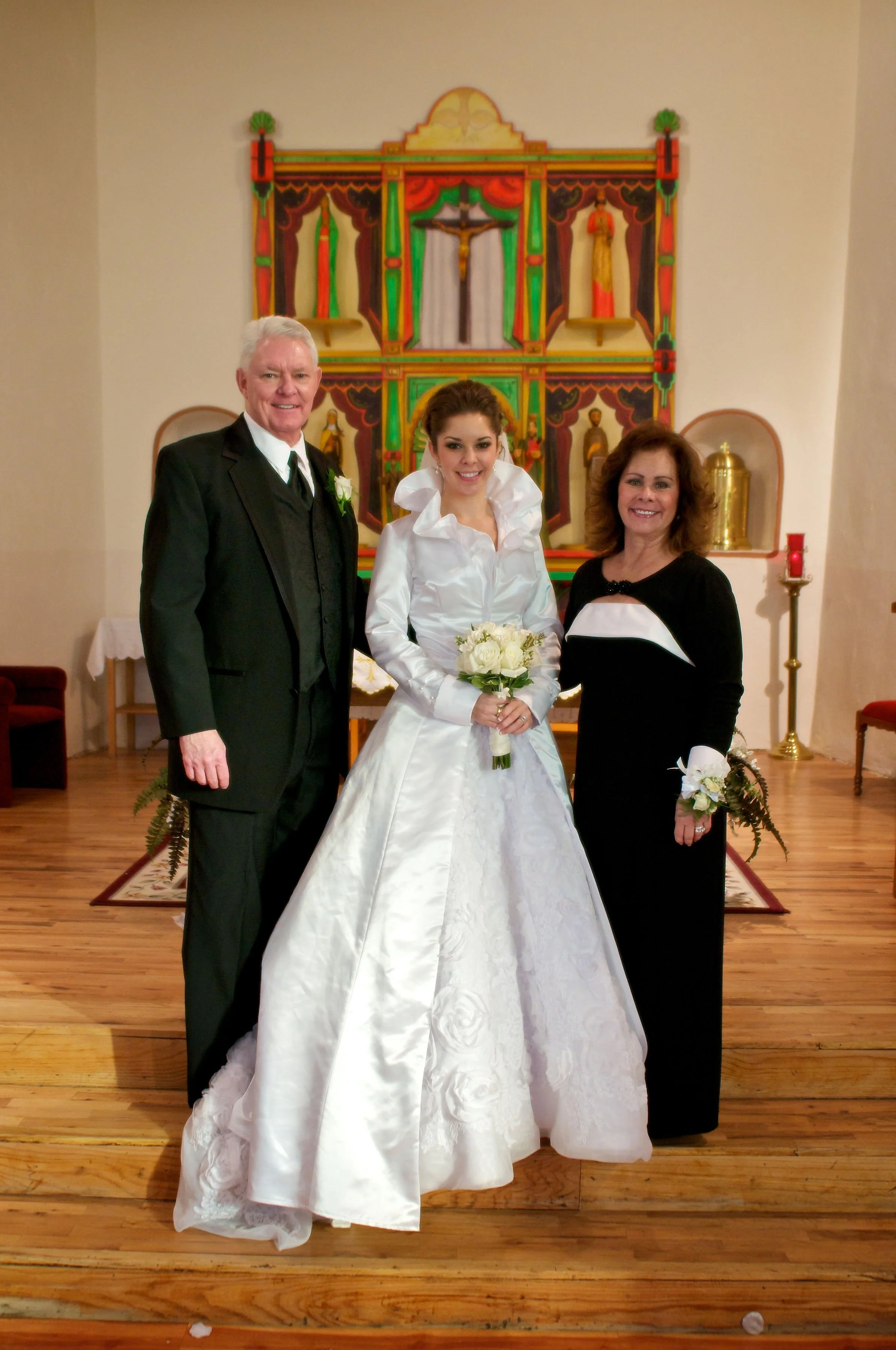 A bride in a white wedding dress holding a bouquet, standing between a man and a woman, inside a church with religious artwork in the background.