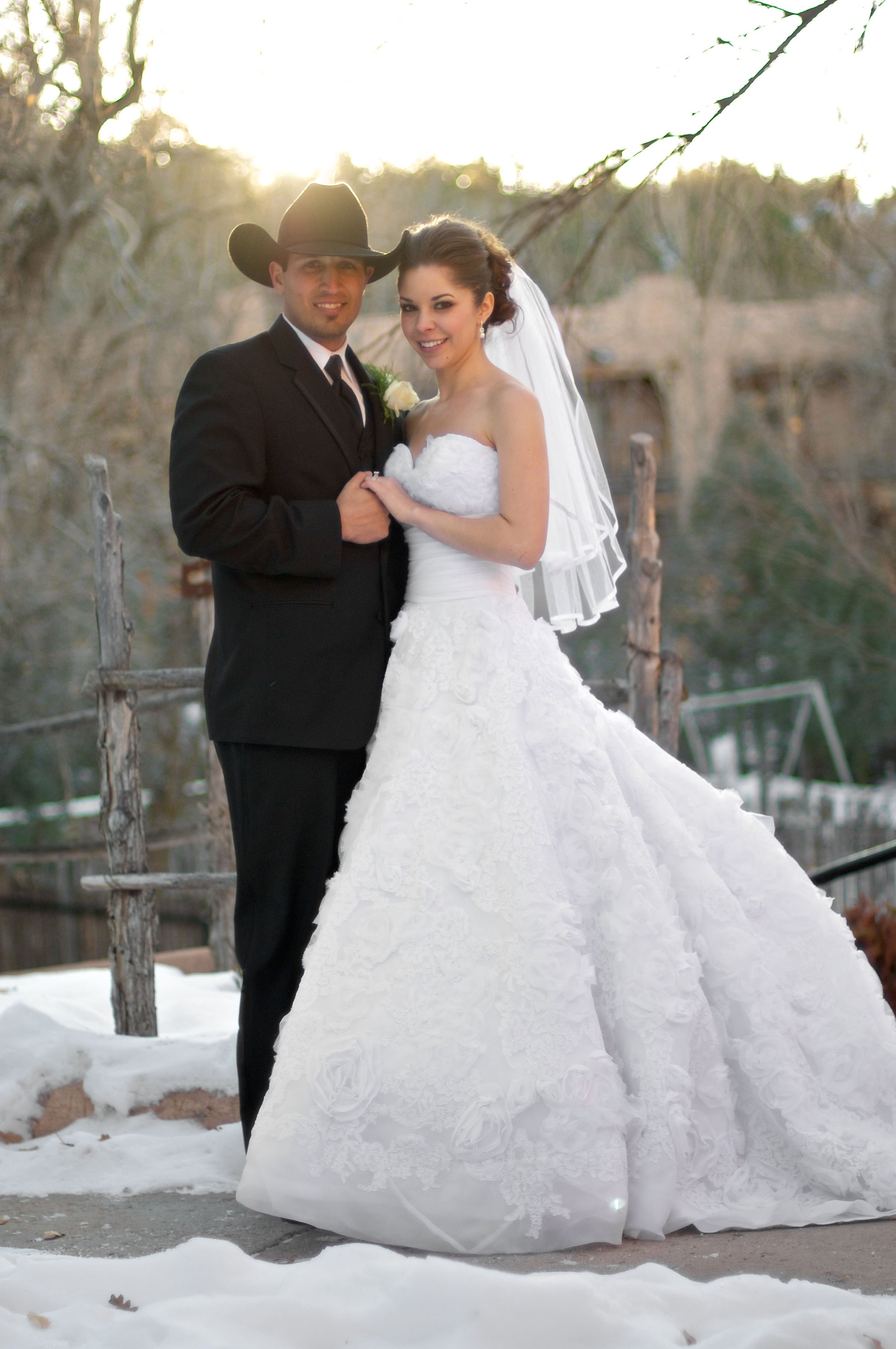 A bride in a white wedding dress and a groom in a black suit and cowboy hat standing outdoors in a snowy area during sunset.