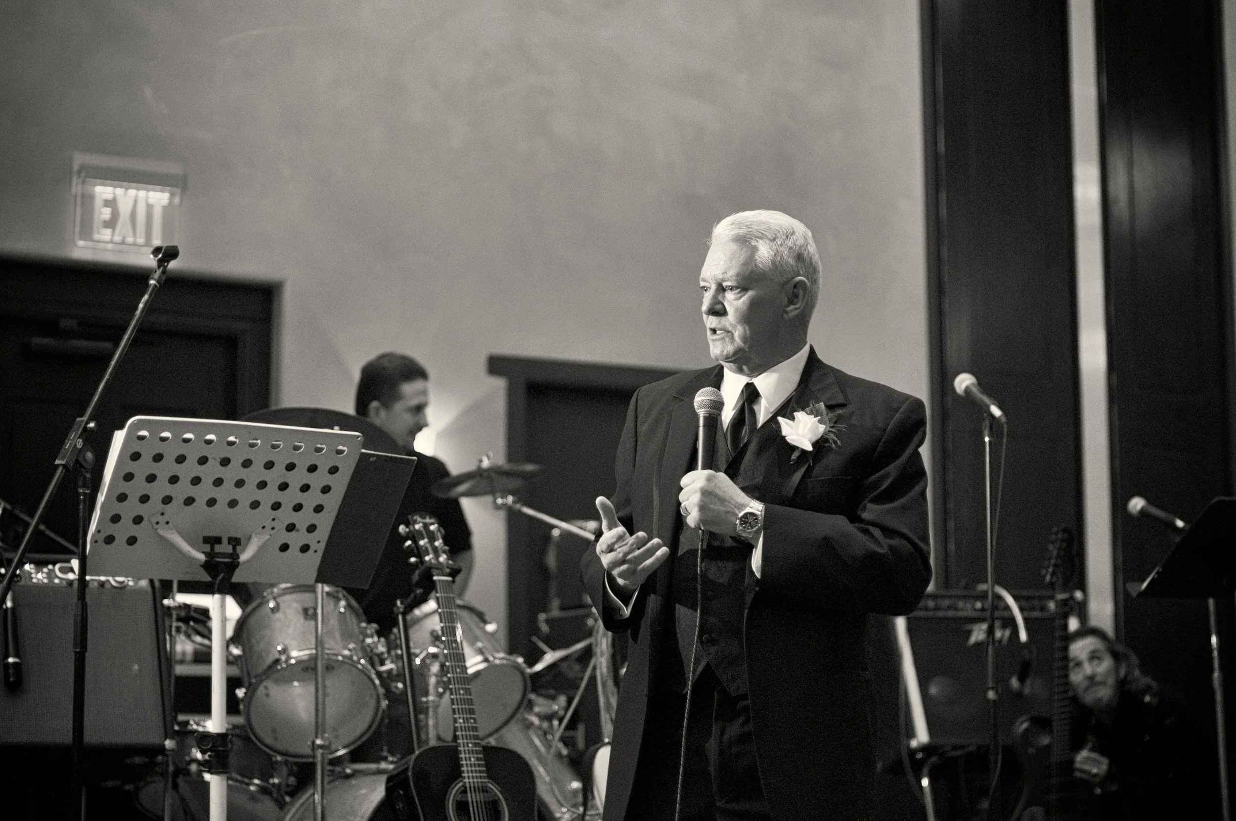 Black and white photo of an elderly man in a suit holding a microphone, standing on a stage with musical instruments and a band in the background.