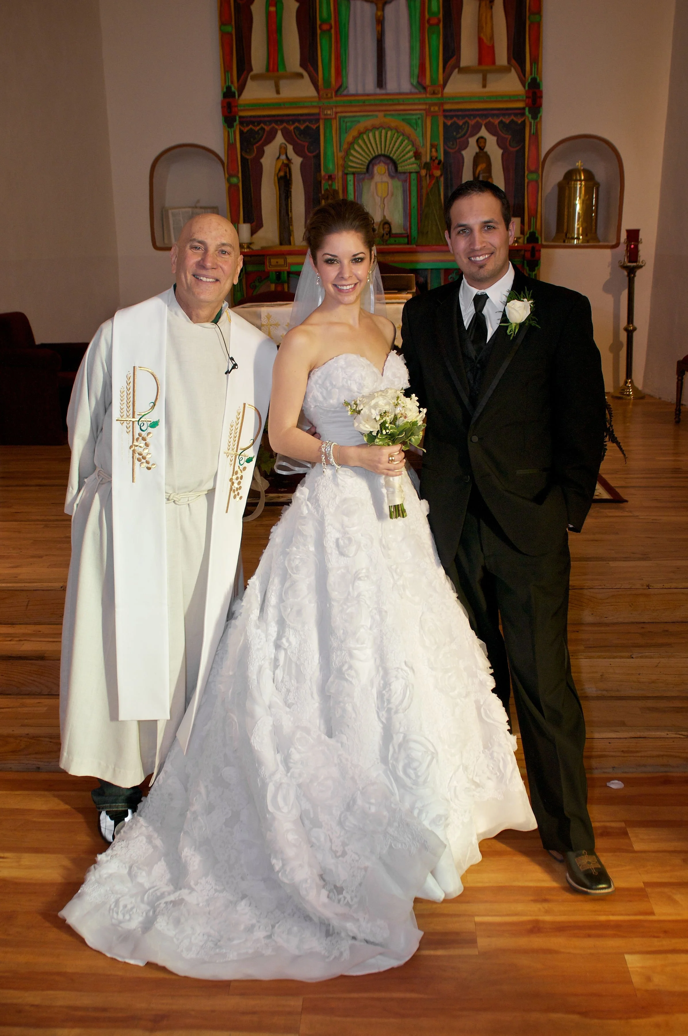A bride and groom standing with a priest in a church after their wedding. The bride is holding a bouquet and wearing a white wedding dress, the groom is in a black suit with a white boutonniere, and the priest is dressed in white robes.