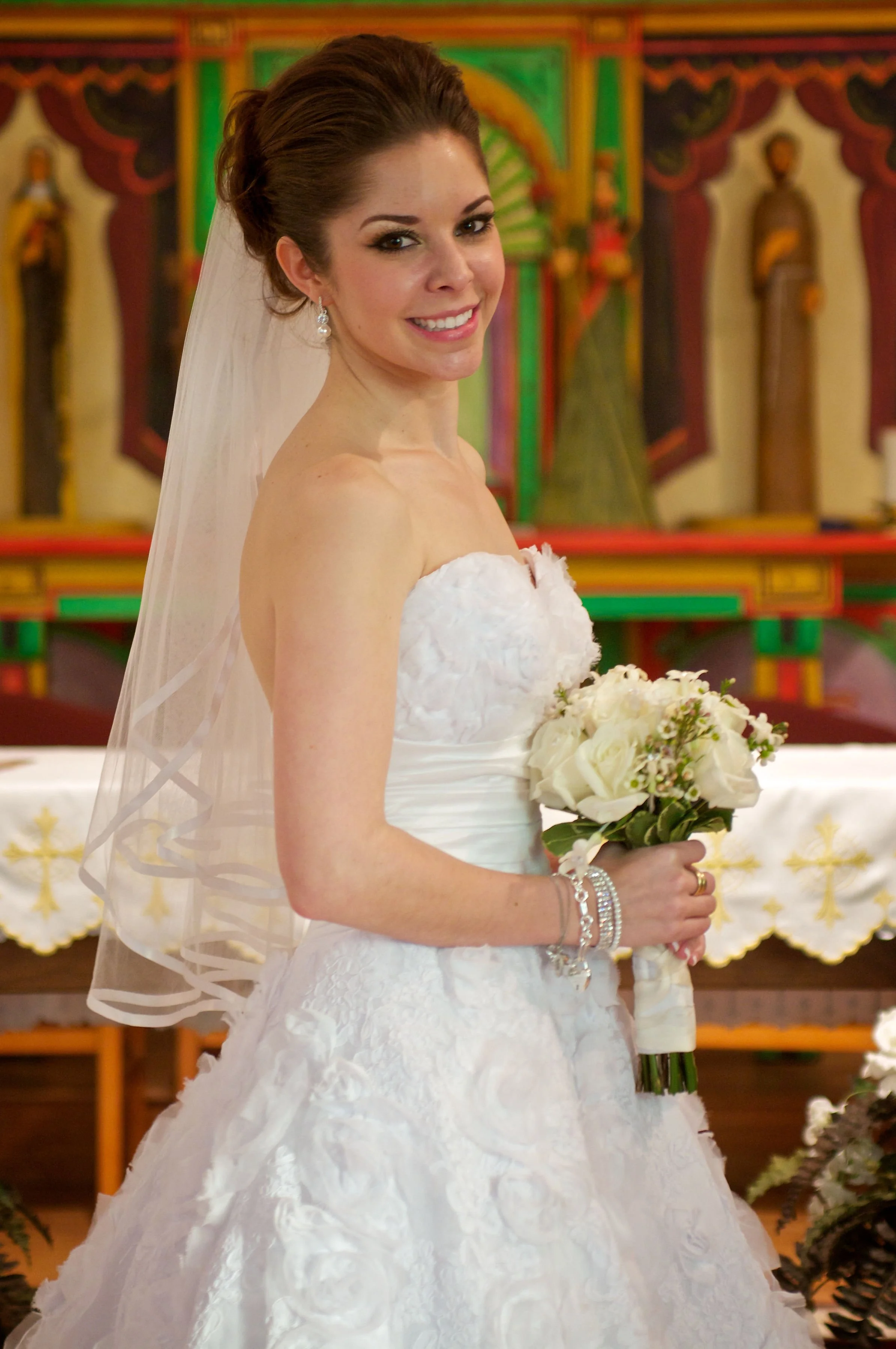 A bride in a white wedding gown holding a bouquet of white roses and greenery inside a church.