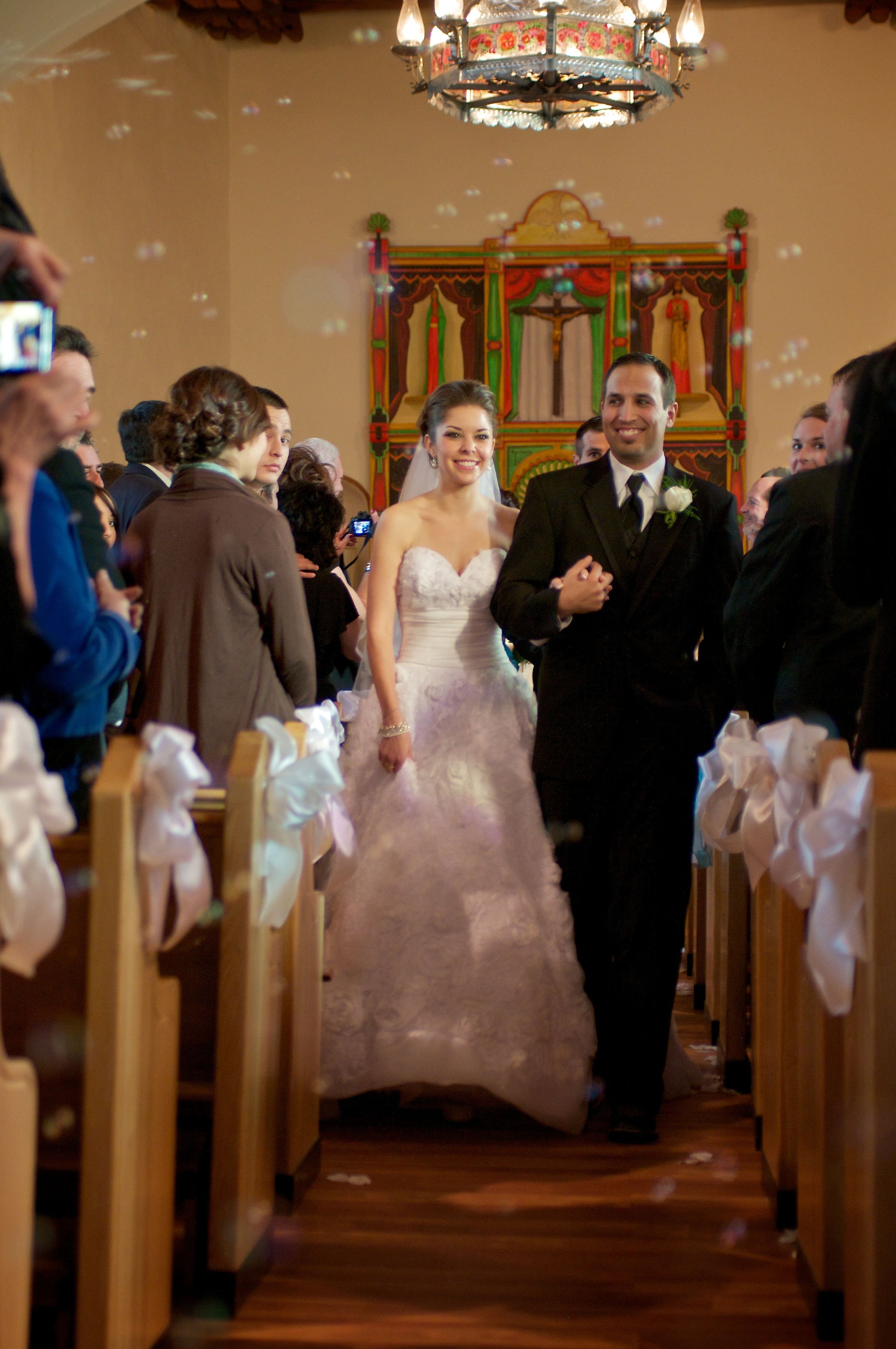 A bride in a white wedding dress and a groom in a black suit walking down the aisle of a church, smiling. Guests on both sides of the aisle watch and take photos. Church decor includes a colorful backdrop, candles, and bow decorations on the pews.