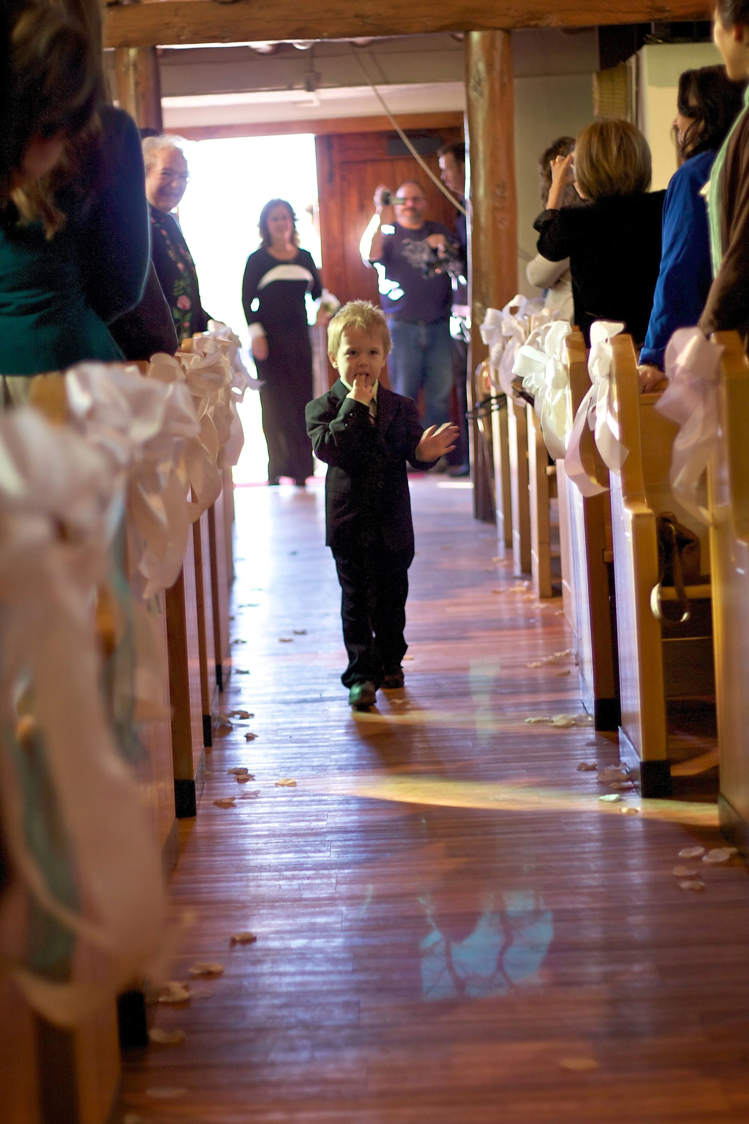 A young boy in a suit walking down an aisle decorated with white ribbons and flower petals, with people standing on either side, at what appears to be a wedding ceremony.