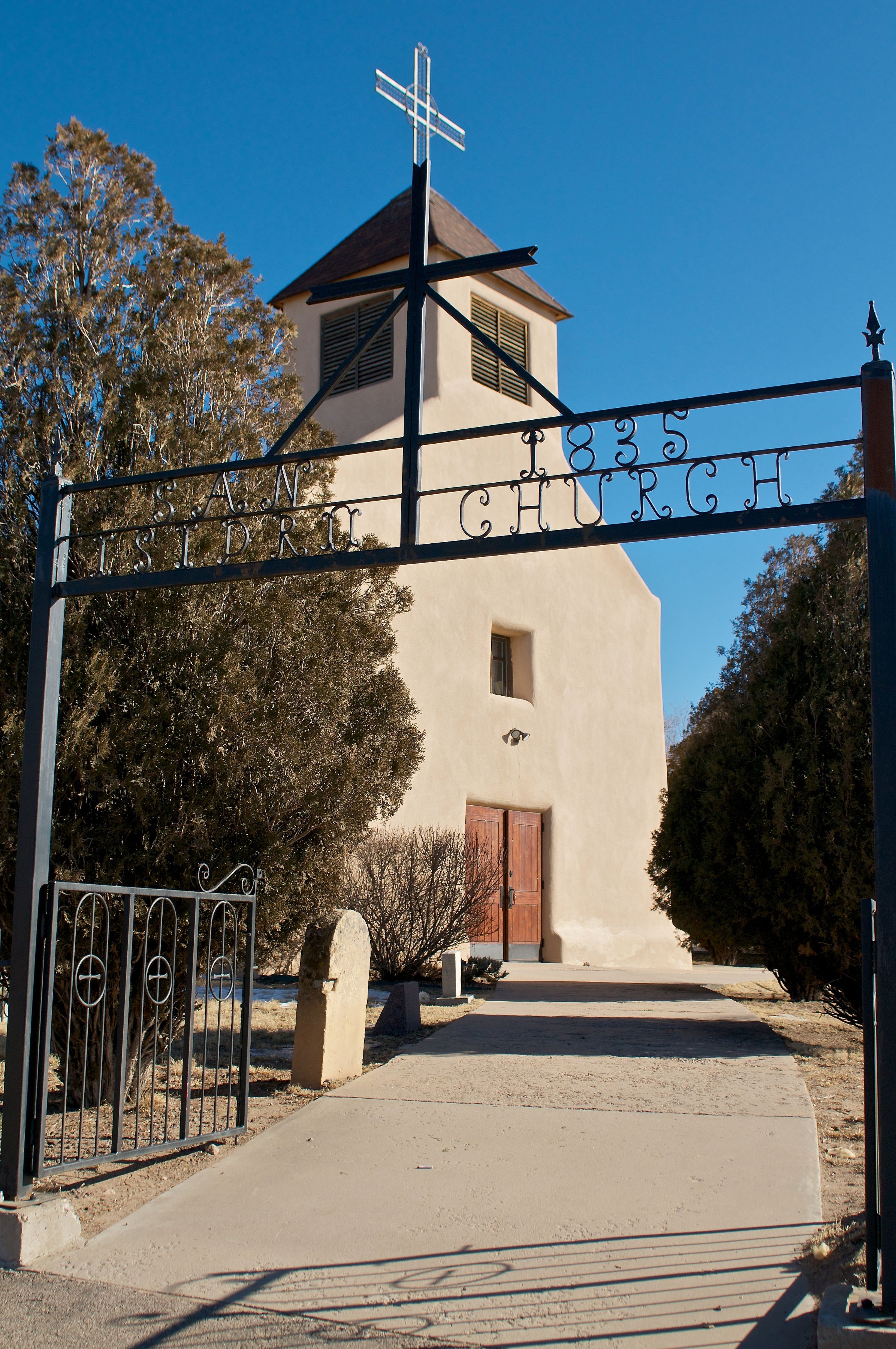 A small, white church with a wooden door and a multiple-sized window, viewed through an iron gate with the words "San Francisco 835 Church." A large cross on top of the church, with trees on either side and a clear blue sky.