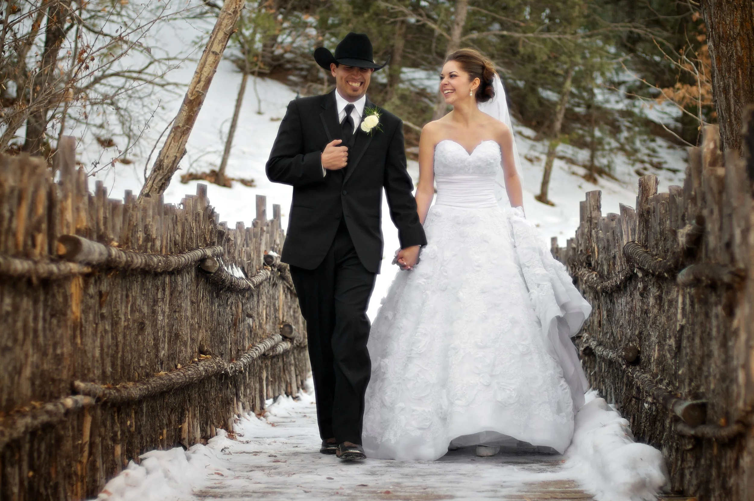 A newlywed couple walking hand in hand on a snowy wooden bridge outdoors, with the bride in a white wedding gown and veil, and the groom in a black suit and cowboy hat, smiling at each other.