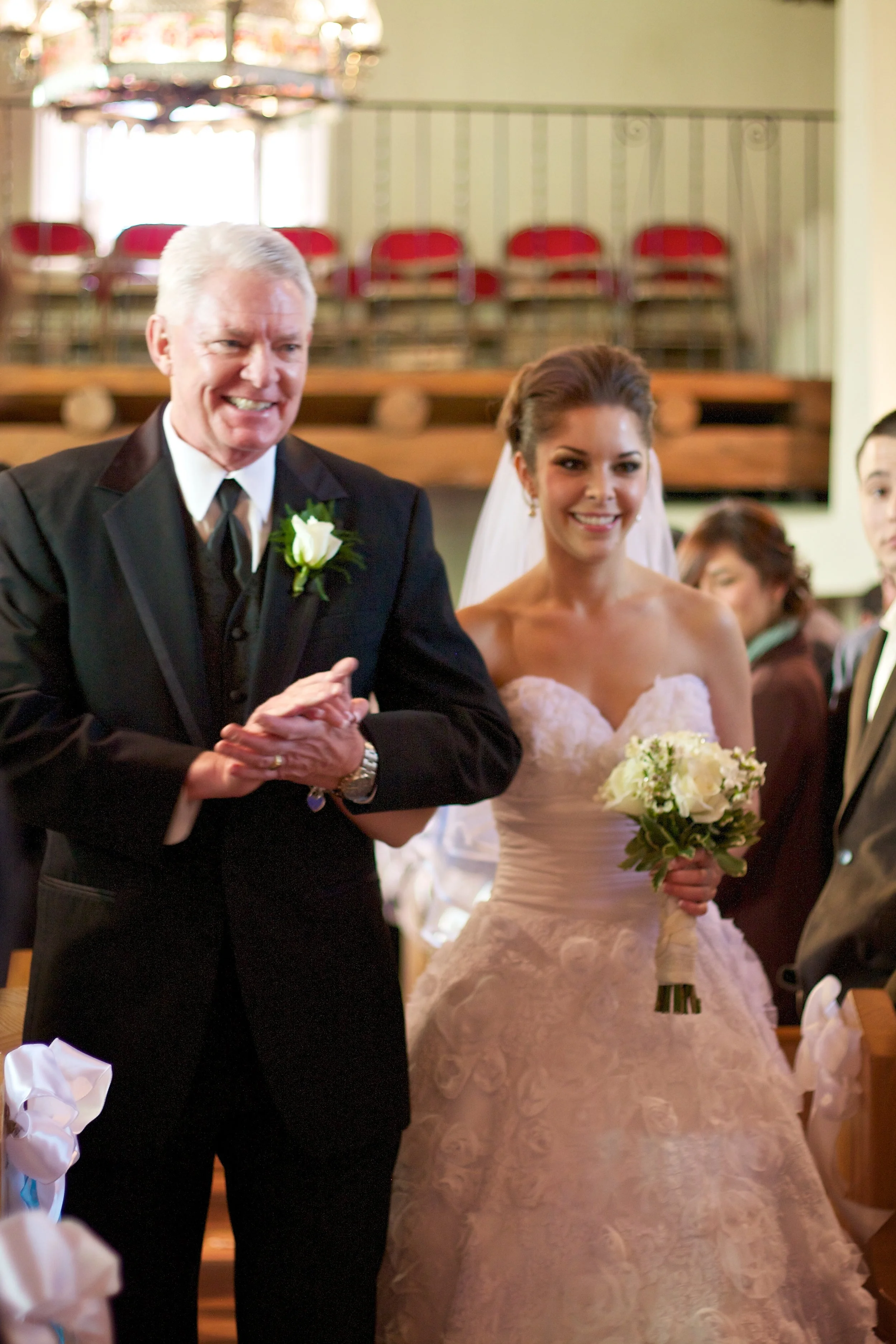 A bride in a white wedding gown holding a bouquet walking down the aisle with an older man, possibly her father, during a wedding ceremony in a church.