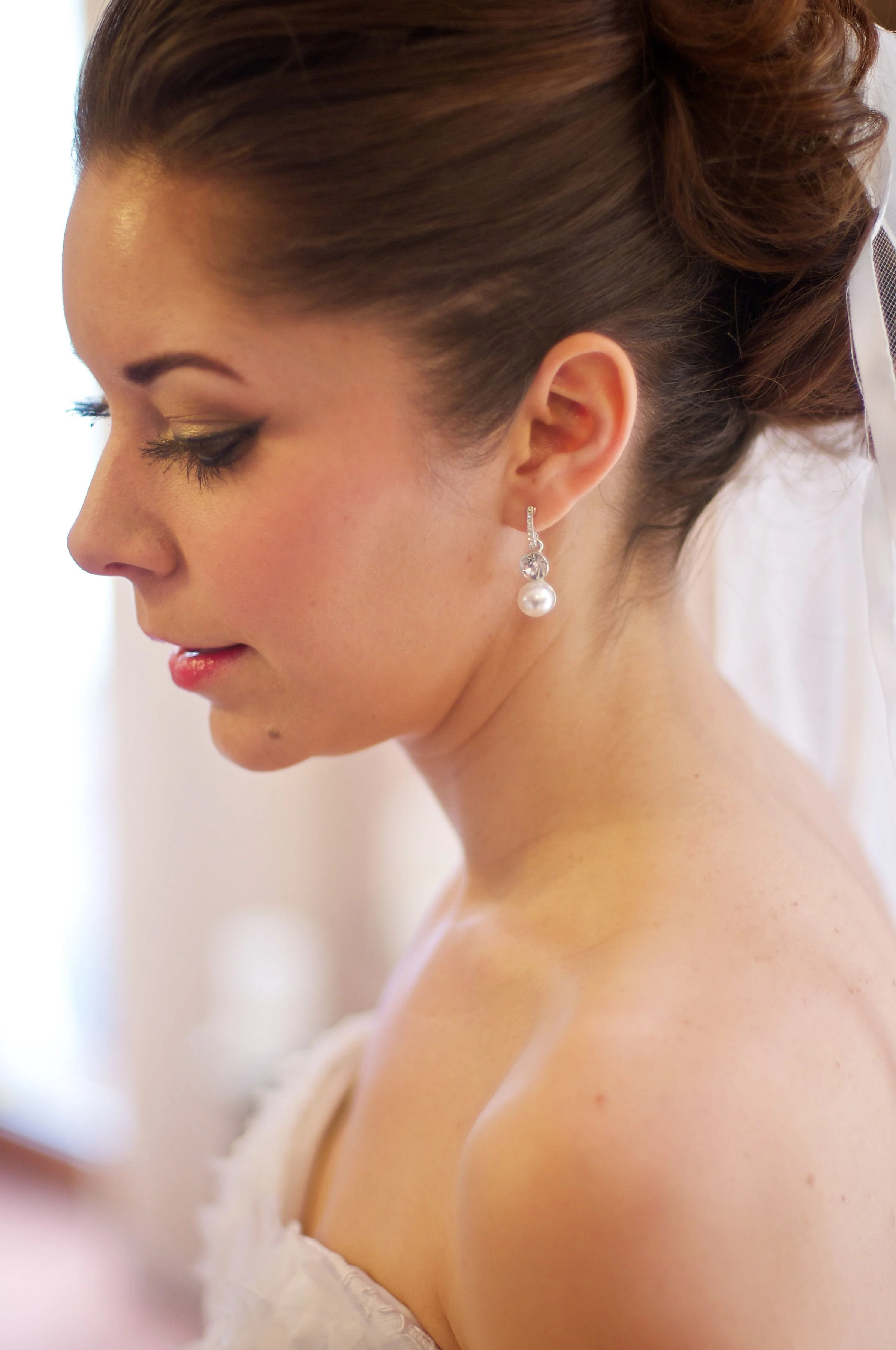 Close-up of a woman with styled brown hair in a bridal updo, wearing pearl and diamond earrings, with makeup on her eyelids, in a softly lit setting.