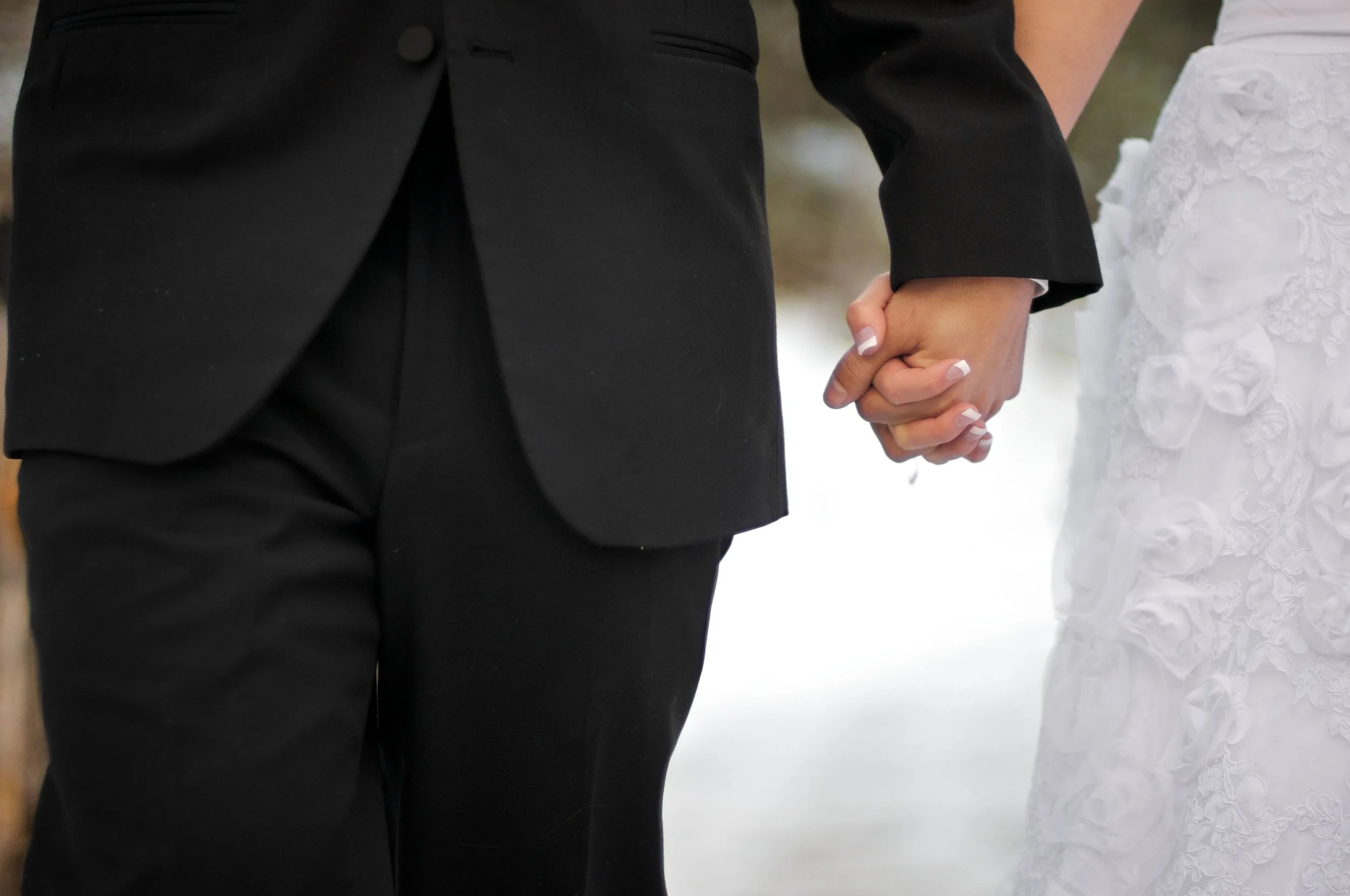 Close-up of a bride and groom holding hands, with the bride wearing a white dress with floral details and the groom in a black suit, outdoors in a snowy setting.
