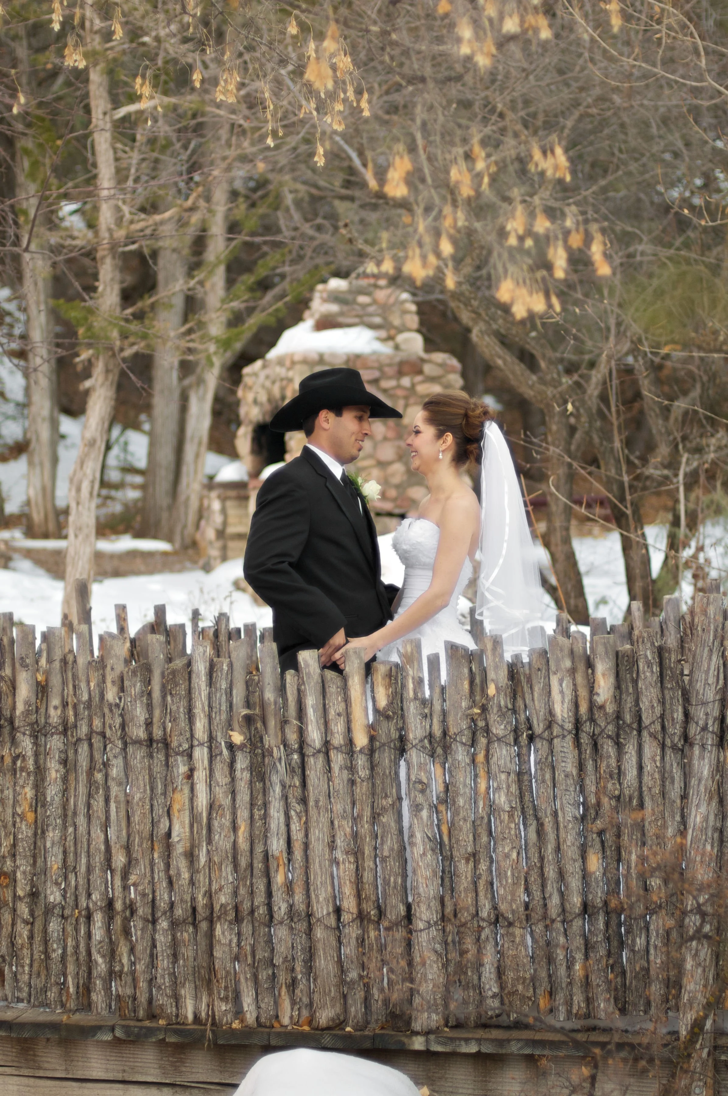 A bride and groom standing behind a rustic wooden fence outdoors, with snowy ground and leafless trees, sharing a romantic moment during their wedding celebration.