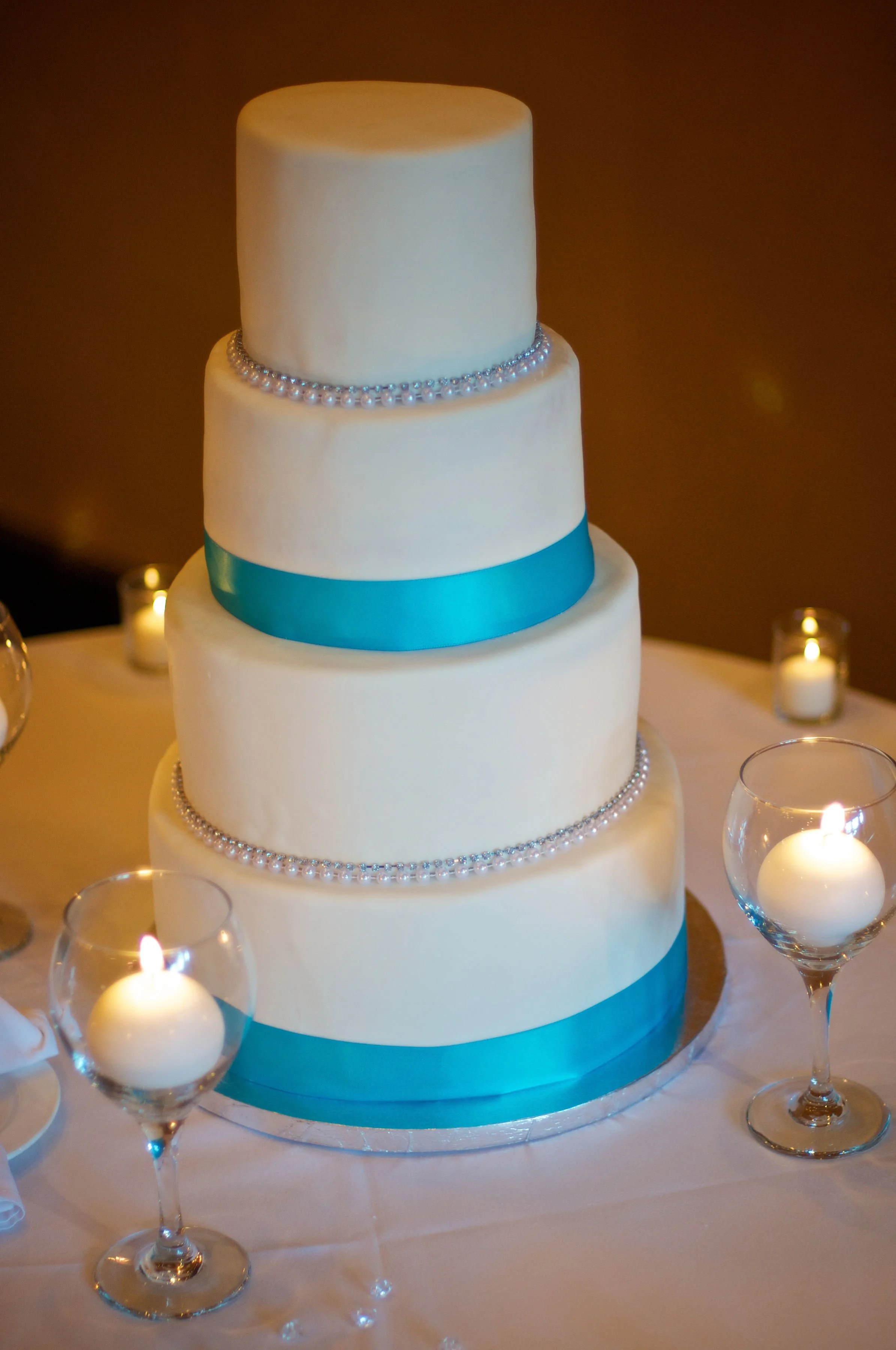 Four-tier white wedding cake decorated with pearls and blue ribbons, surrounded by lit candles in glass holders.