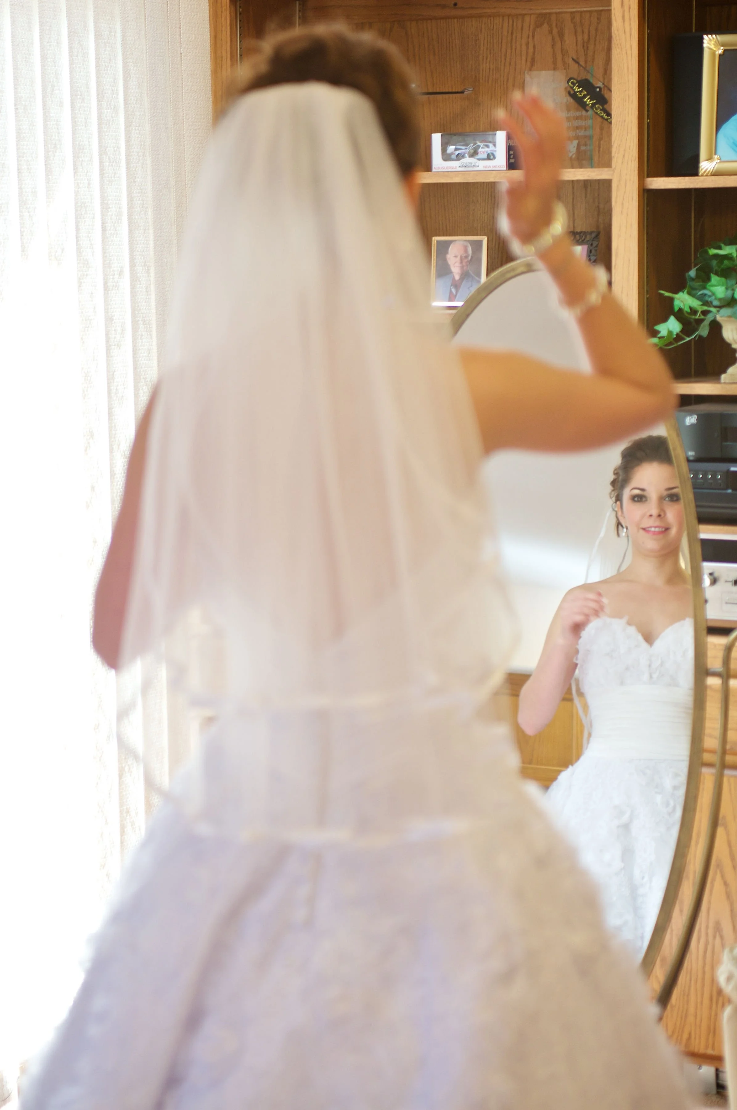 A bride dressed in a white wedding gown and veil looking at her reflection in a mirror, adjusting her hair.