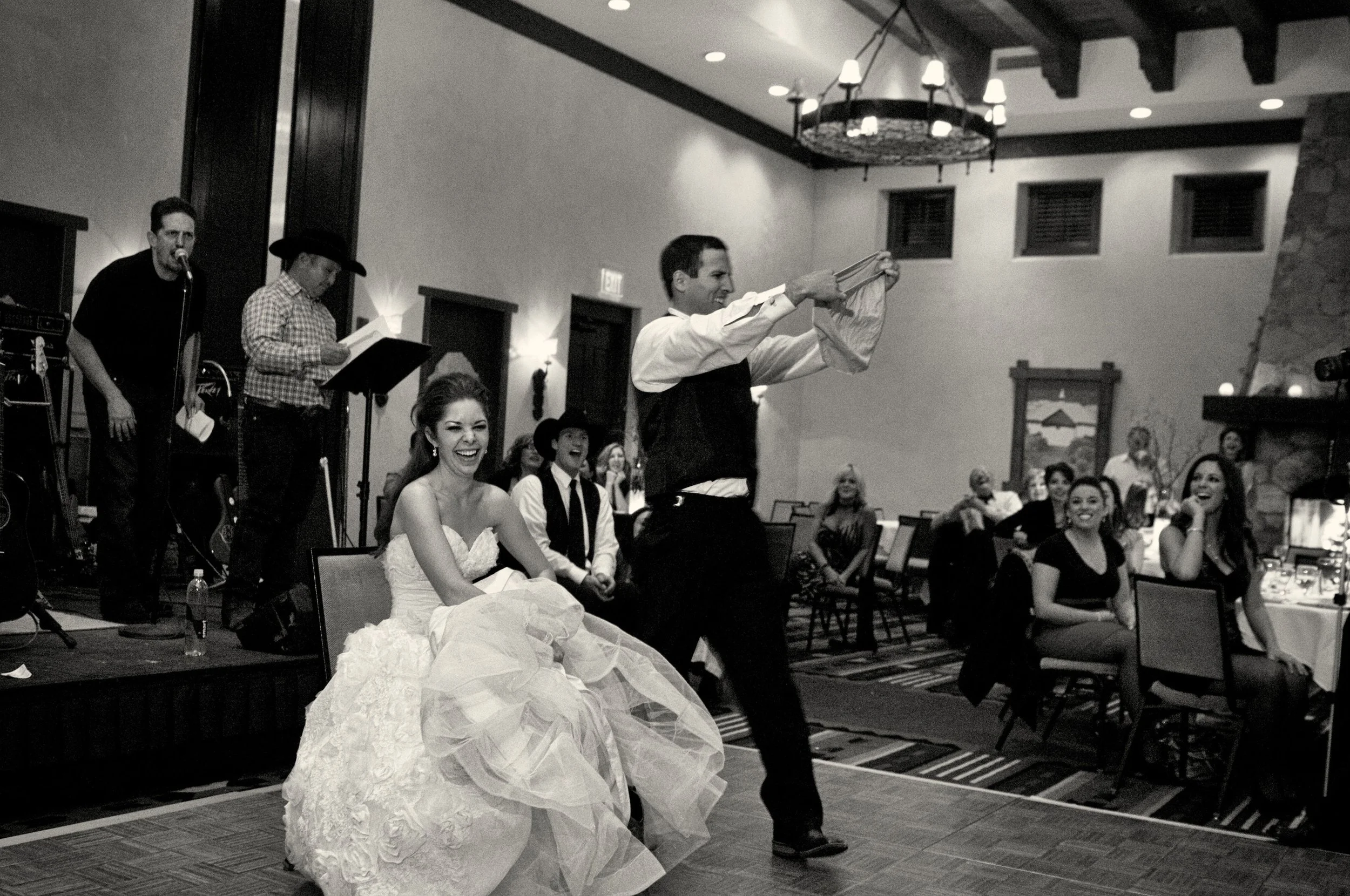 Bride in a strapless wedding gown laughing while sitting on a chair and groom in black vest and white shirt holding a folded cloth during a wedding reception in a spacious hall with guests seated at tables.