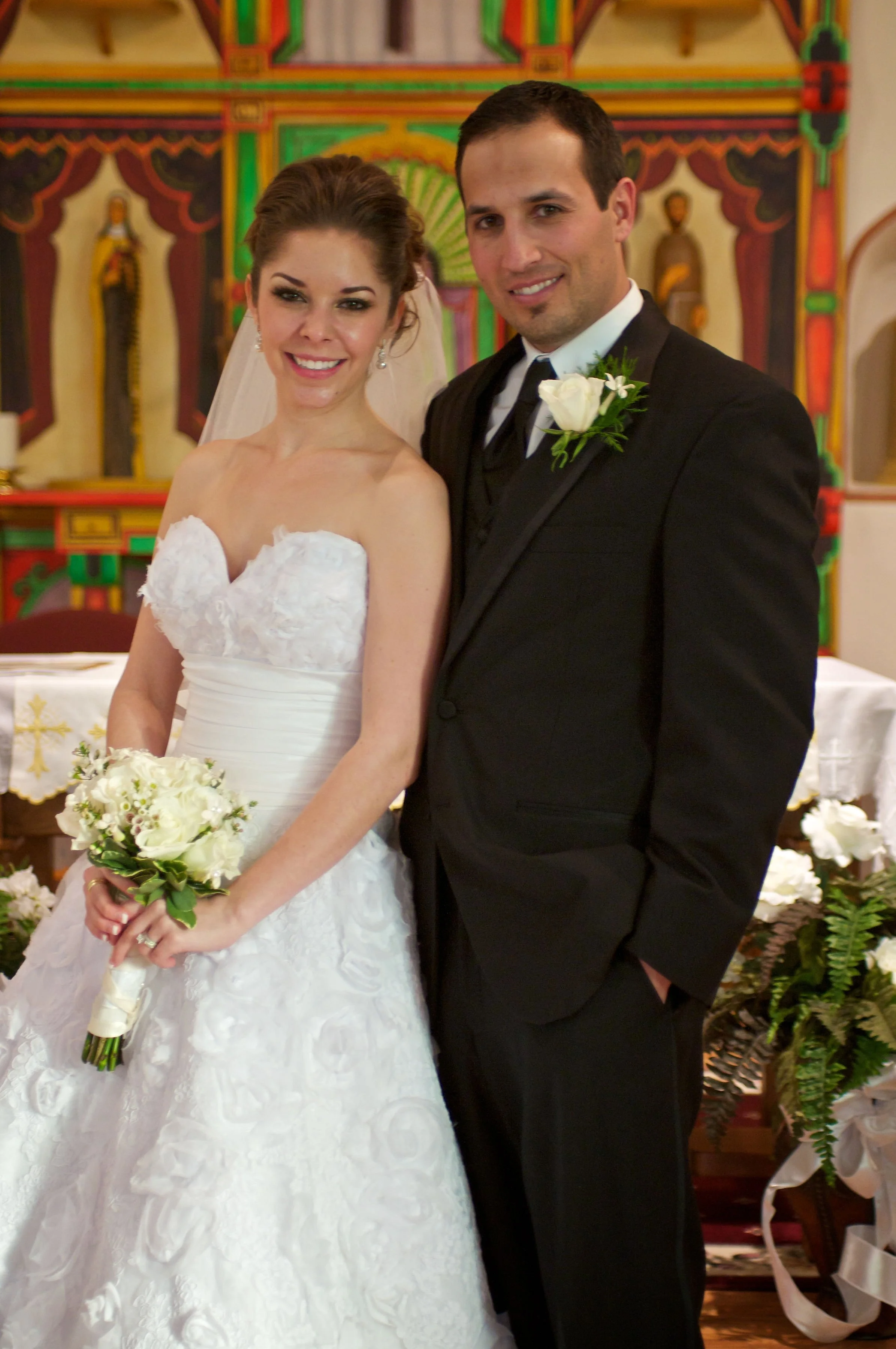 A bride and groom at their wedding ceremony, standing together inside a church, with the bride holding a bouquet of white flowers and the groom wearing a black tuxedo with a white boutonniere.