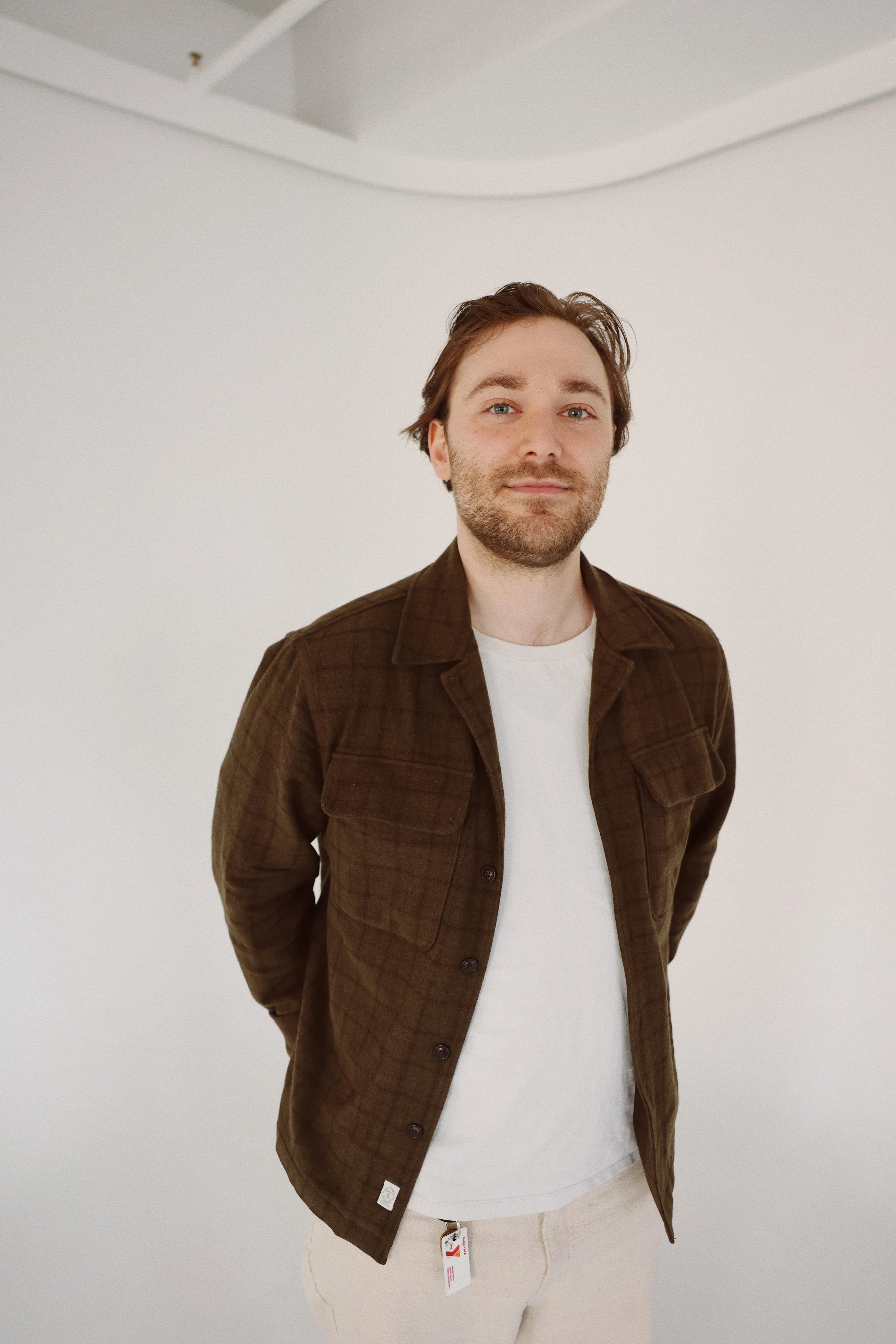 A man with brown hair and a beard standing against a plain white background, wearing a brown plaid jacket over a white t-shirt and beige pants.