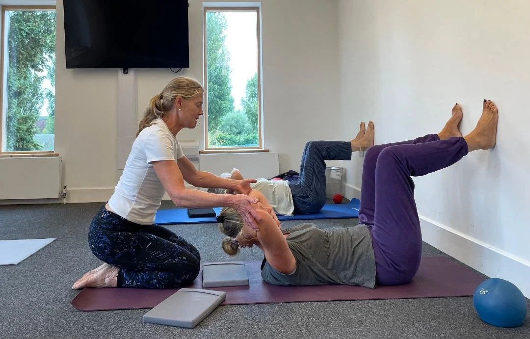 A woman assisting an elderly woman with stretching exercises on a yoga mat, with another person doing stretches in the background in a bright room.