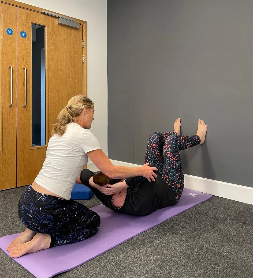 A woman assisting another woman with an exercise on a purple yoga mat in a room with gray walls and wooden doors. The woman being assisted is lying on her side with her legs bent and feet against the wall, while the woman assisting is holding her upper body.