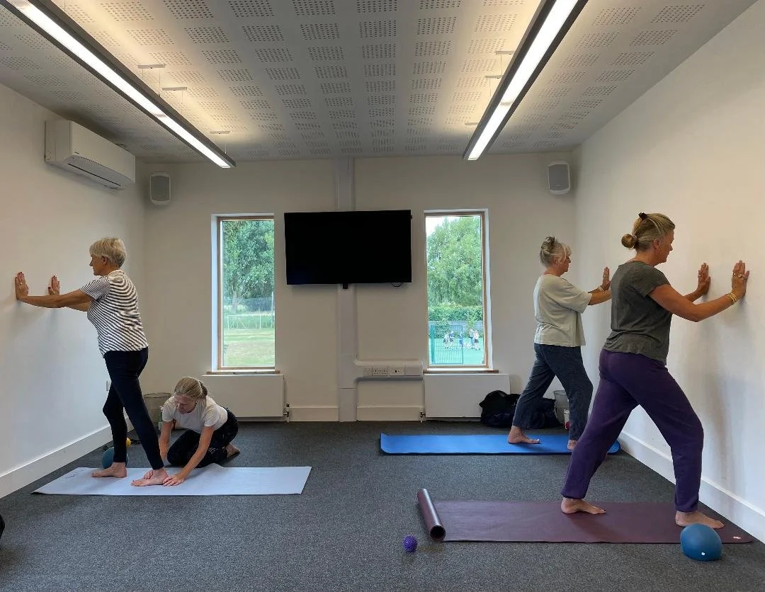 Four women practicing yoga in a bright room with large windows, two on adjustable mats, doing stretches against the wall, with yoga props on the floor.