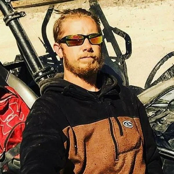 A man with red hair, sunglasses, a beard, and a black and brown jacket standing in front of an off-road vehicle.