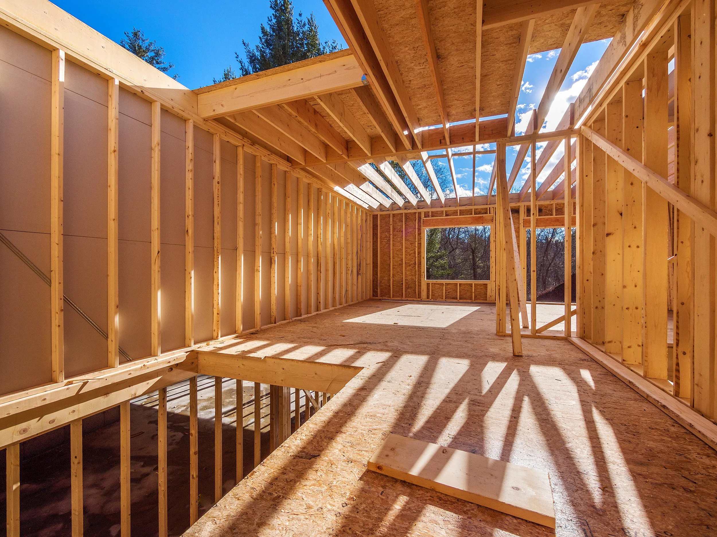 Inside a wooden house under construction, showing framing with sunlight casting shadows through the open roof and window.