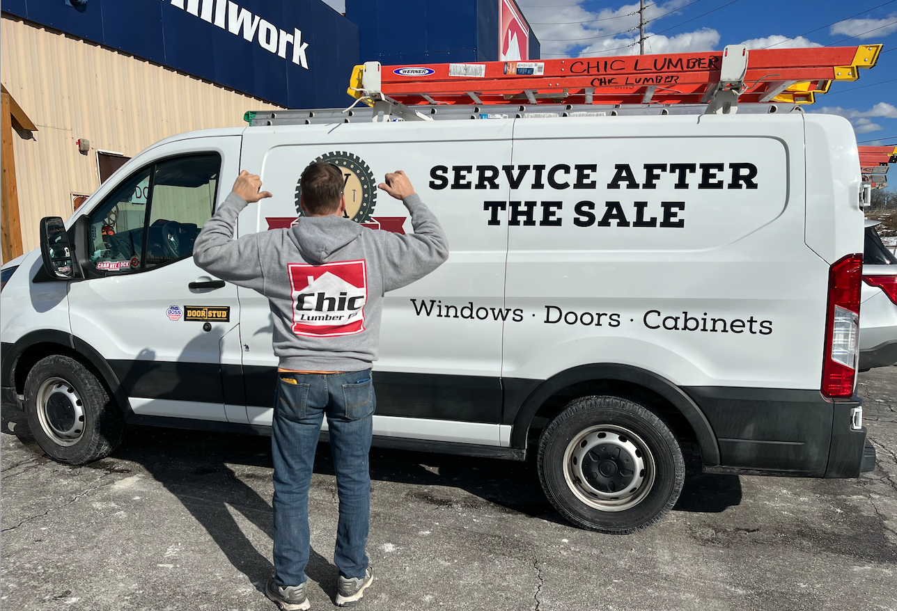 A man in a gray hoodie with a Chic Lumber logo on the back, standing in front of a white service van with the words "SERVICE AFTER THE SALE" and "Windows, Doors, Cabinets" written on the side. The van has a ladder on top labeled "Chic Lumber" and is parked outside a building with a blue sign. The man is flexing his arms, showing his muscles, with his back to the camera.