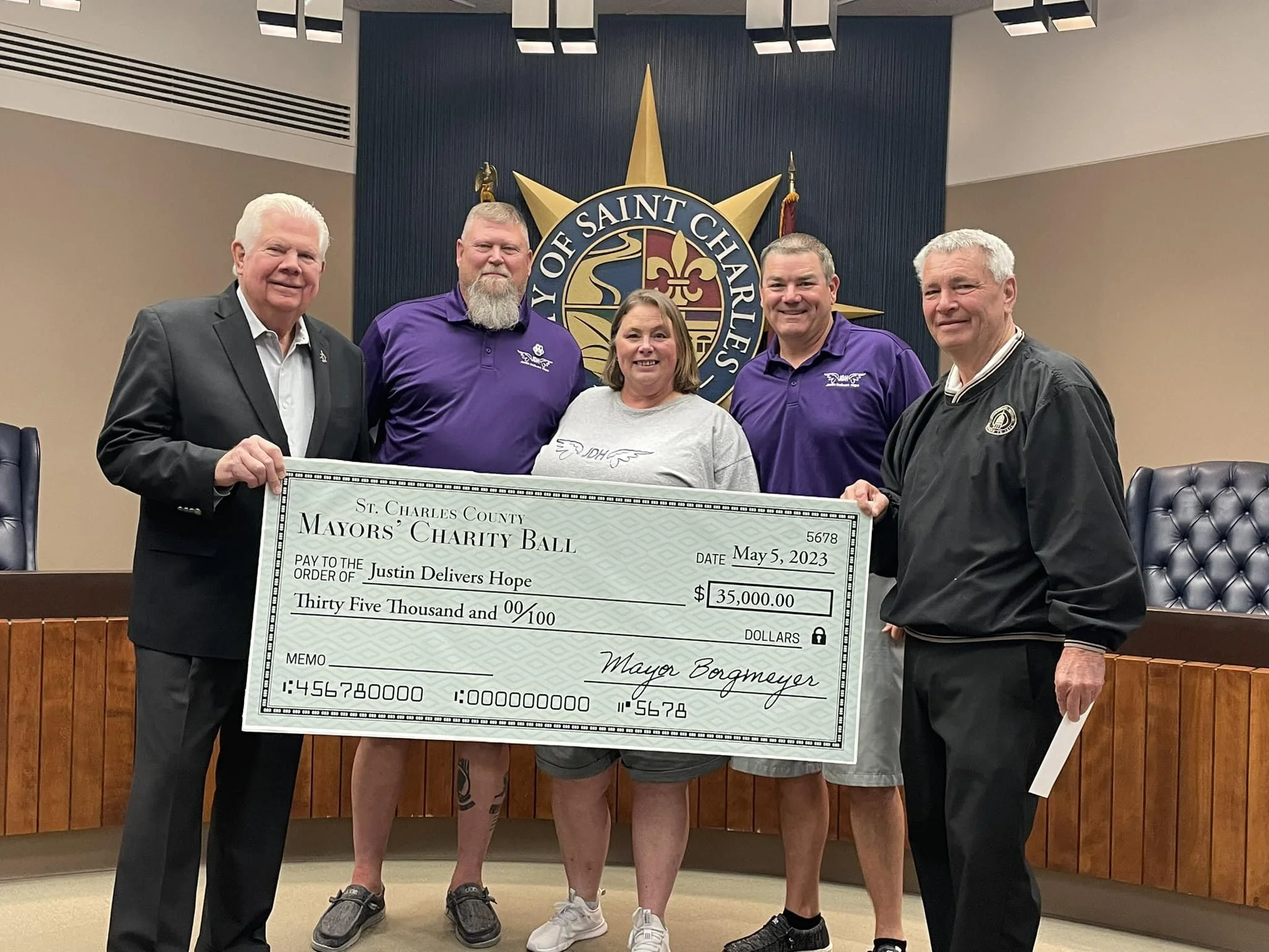 Group of five people standing behind a large check for $35,000 made out to Justin Delivers Hope, at a formal event in a government room with a seal on the wall behind them.