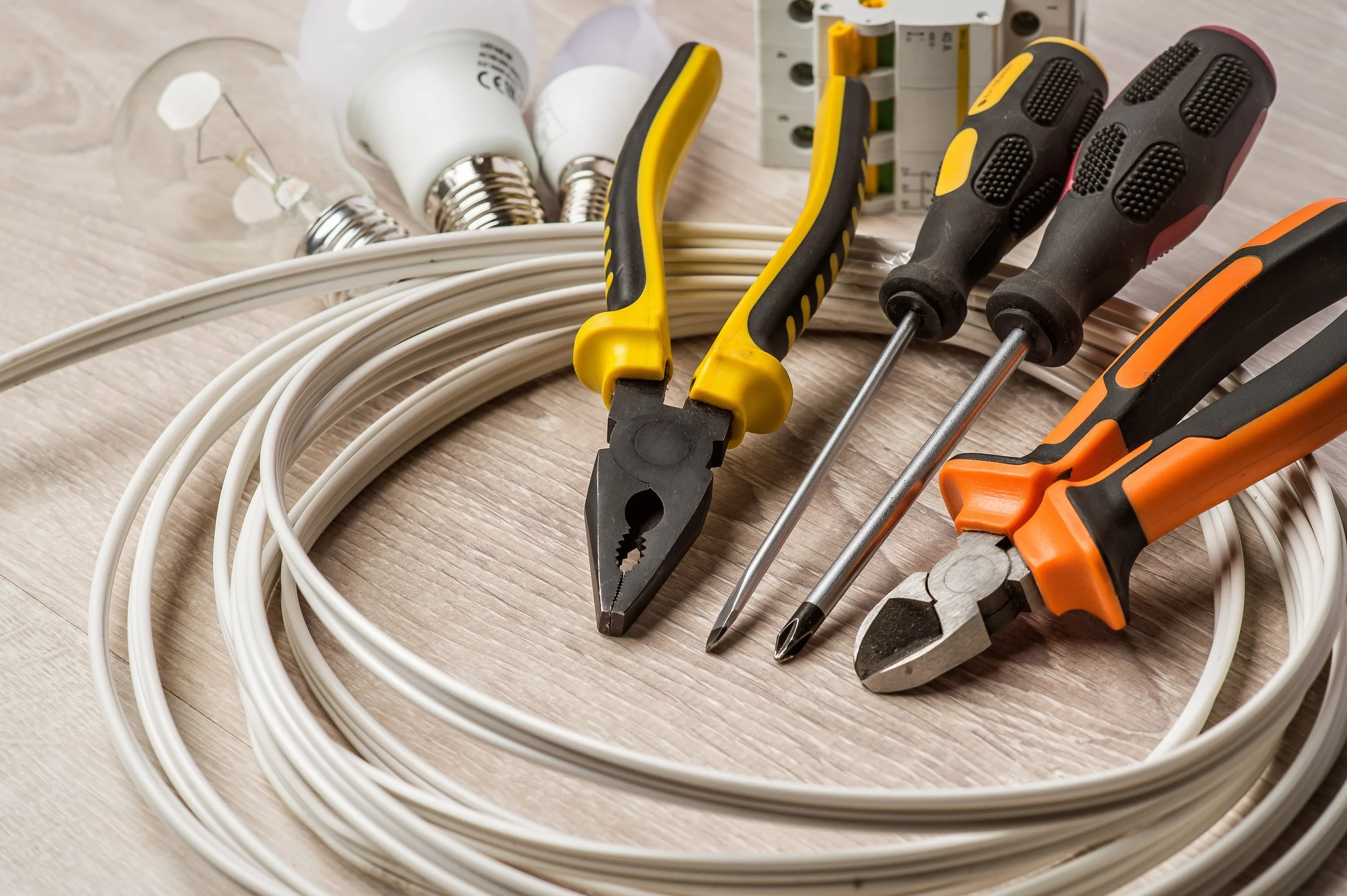 Electrical tools including pliers, screwdrivers, lightbulbs, wiring, and a circuit breaker on a wooden surface.