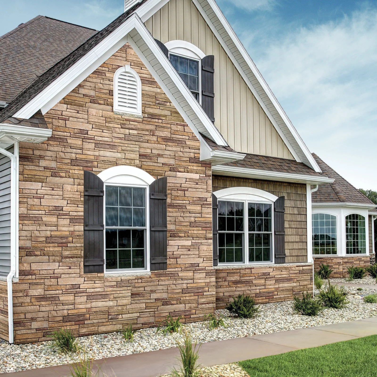 Exterior of a house featuring a combination of brick, siding, and shingle siding, with large windows and black shutters, and a landscaped yard with small bushes and rocks.