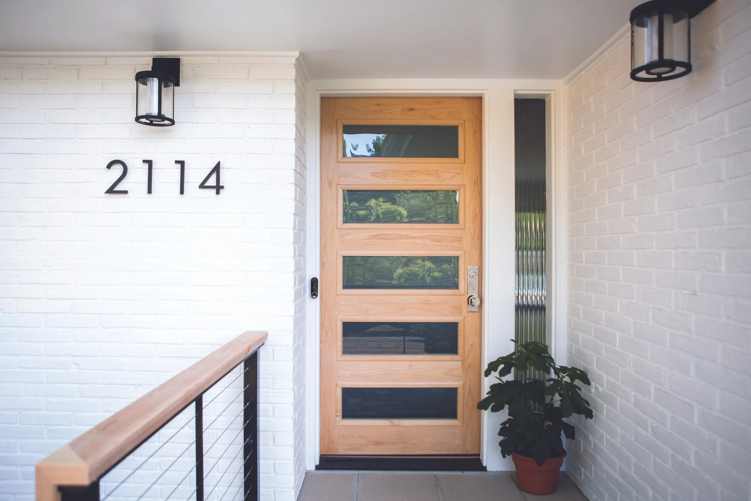 Front porch with a wooden door with horizontal glass panels, house number 2114 on the white brick wall, black wall-mounted lights, and a potted plant on the right side.