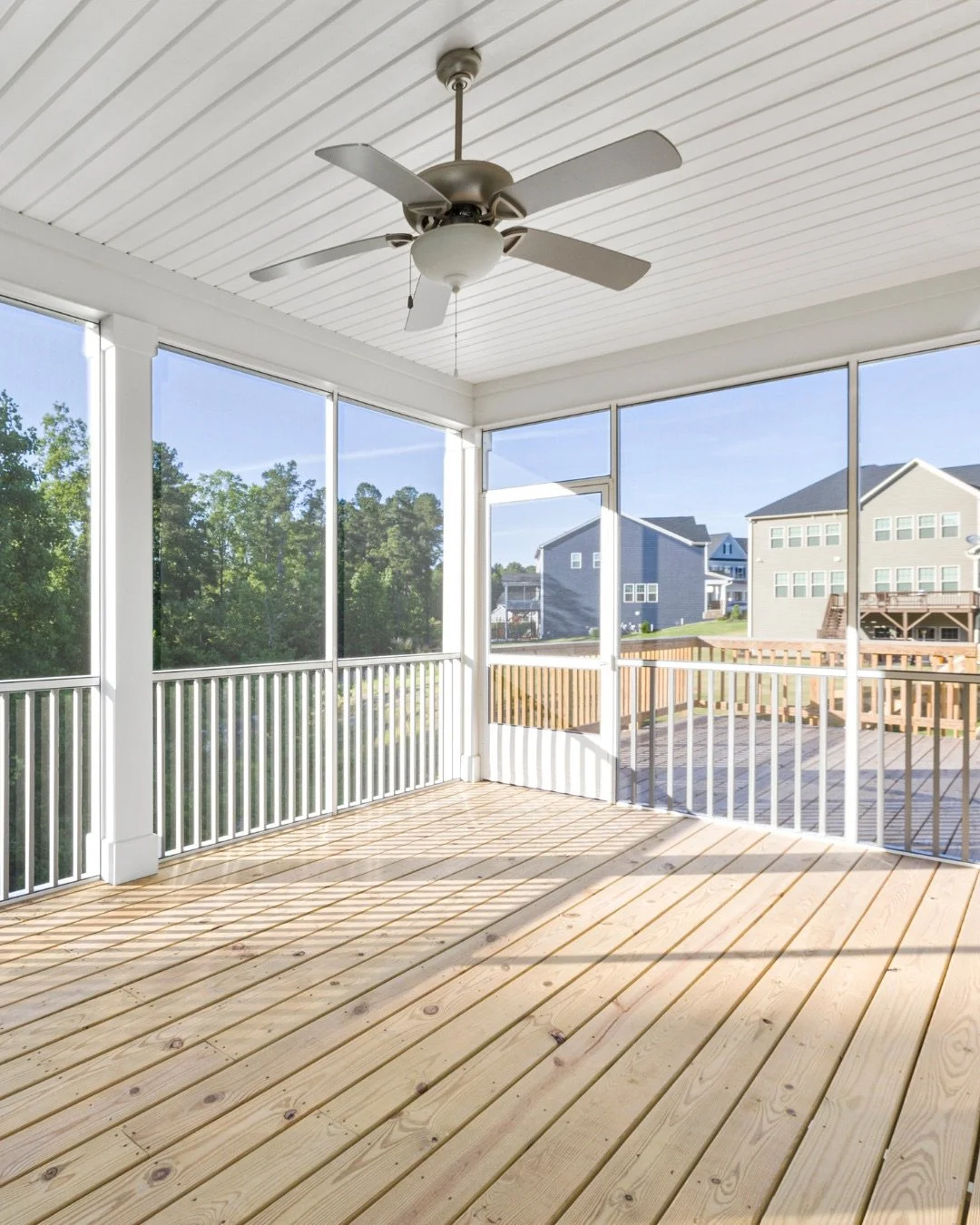 Enclosed porch with natural wood flooring, white railing, ceiling fan, and large windows showing neighboring houses and greenery.